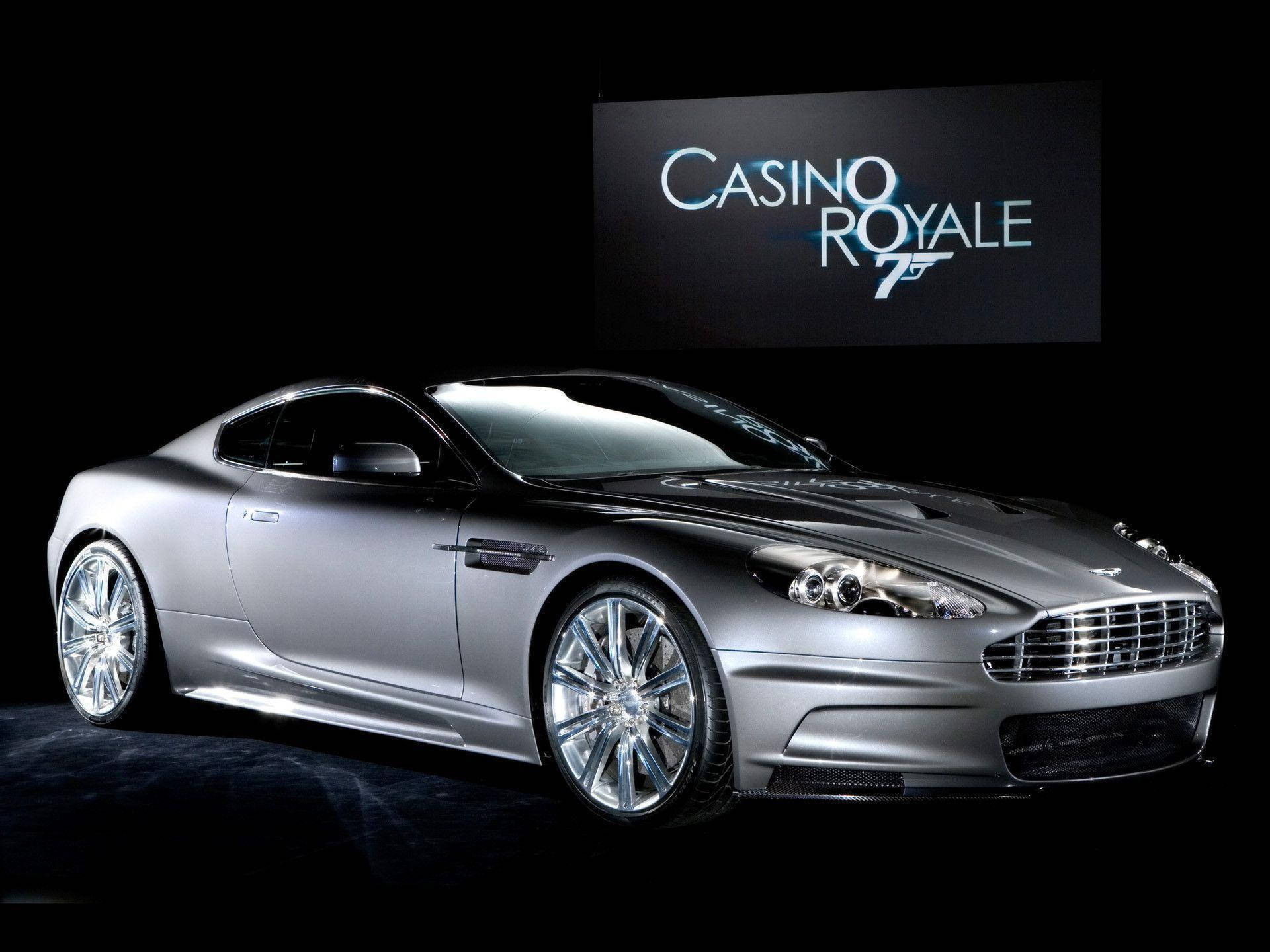 007：皇家賭場 豪華跑車 壁紙 — 一輛時尚的銀色Aston Martin DBS跑車在攝影棚燈光下閃耀，黑色背景襯托著Casino Royale 75標誌屏幕。