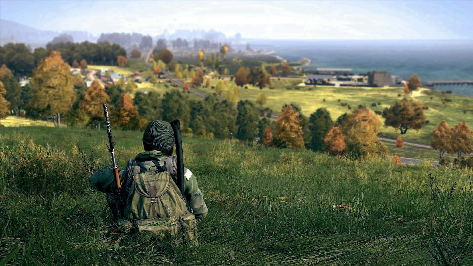 1080p DayZ遊戲背景：男子置身茂密田野 壁紙 — 一名身穿迷彩裝備的士兵坐在長滿草的山丘上，俯瞰著一個擁有秋樹和遠處水域的沿海村莊，天空朦朧。