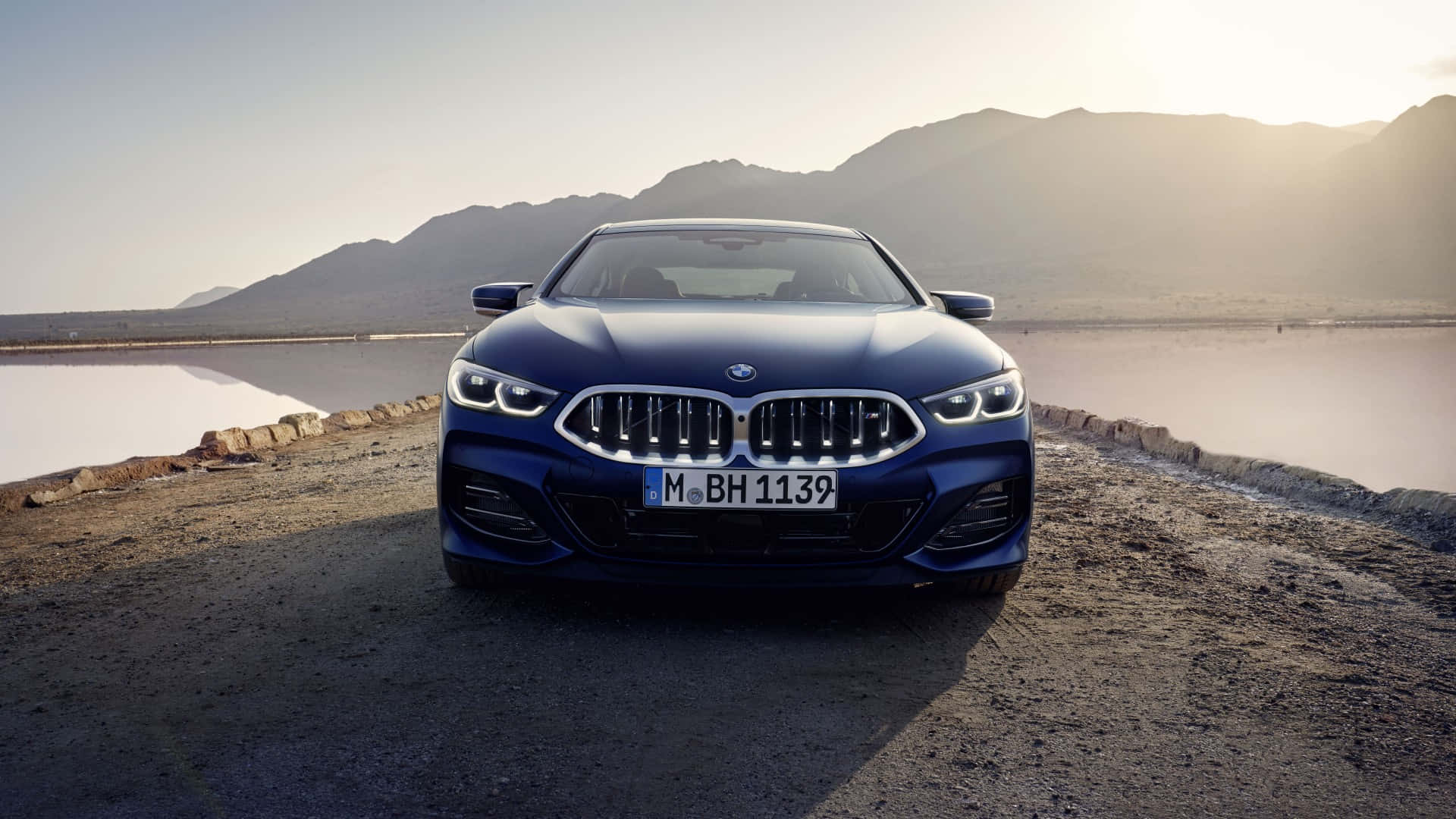 1080p M系列背景：藍色BMW 8系 壁紙 — 一架時尚的藍色寶馬8系轎跑車停在崎嶇的道路旁，旁邊是鹽灘，遠山在朦朧的金色光線中。