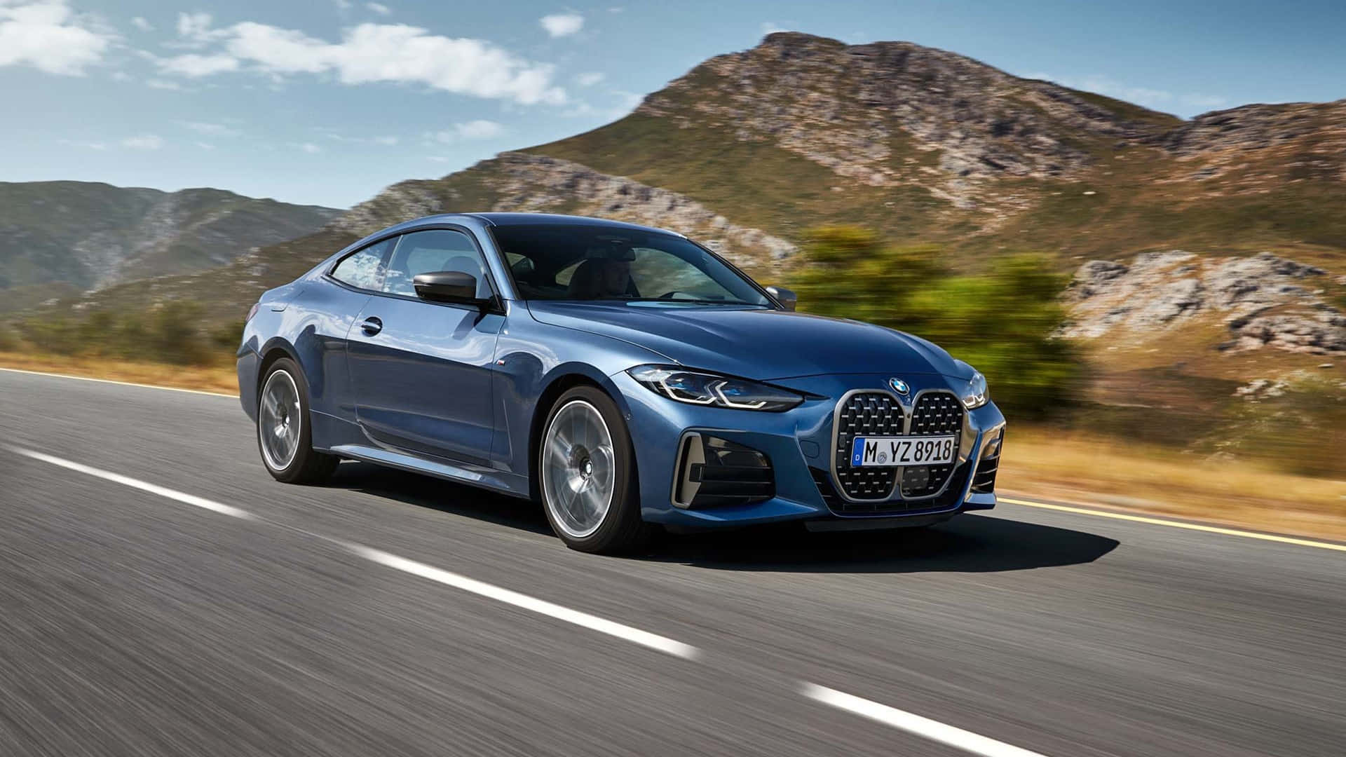1080p M系列背景：2021款灰藍色BMW 4系 壁紙 — 一輛時尚的藍色BMW雙門跑車在晴朗的天空下沿著蜿蜒的山路飛馳。