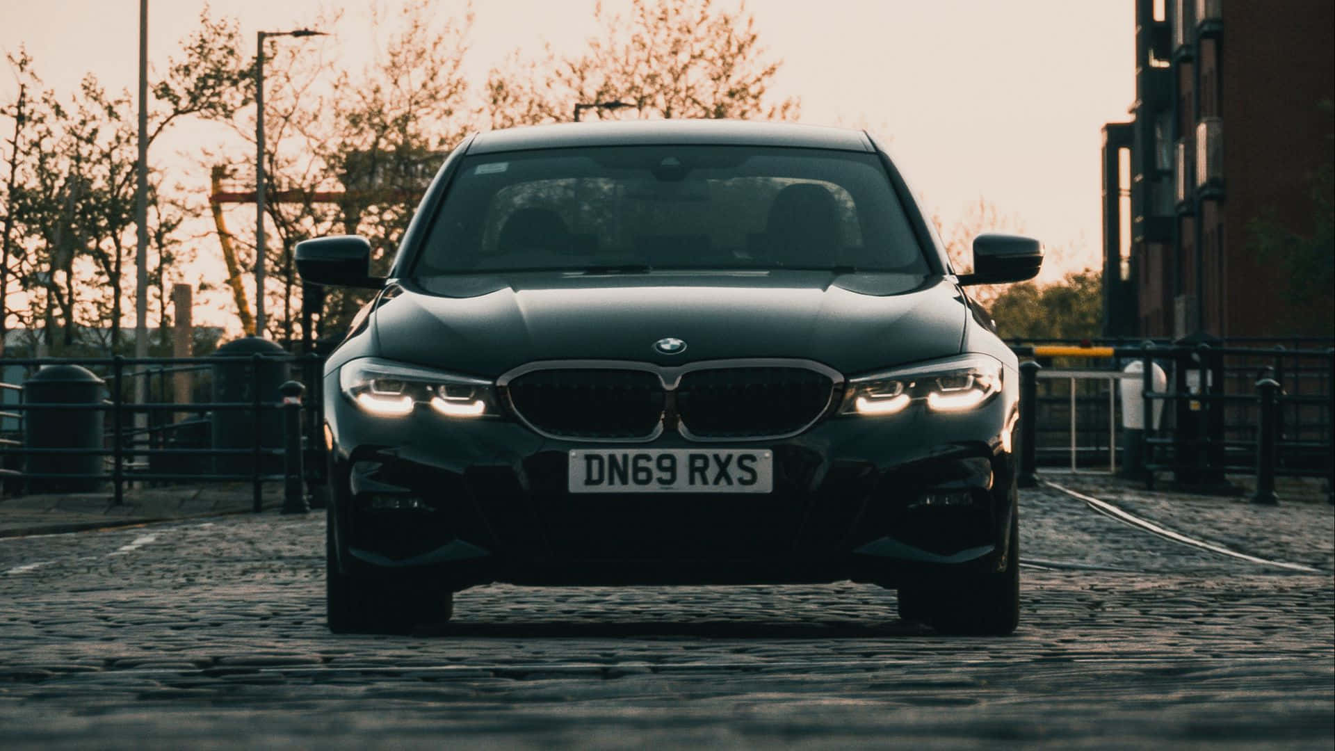 1080p M系列背景：深黑BMW 3系 壁紙 — 一輛時尚的黑色BMW轎車在黃金時段於鵝卵石街道上亮著車頭燈向前行駛。