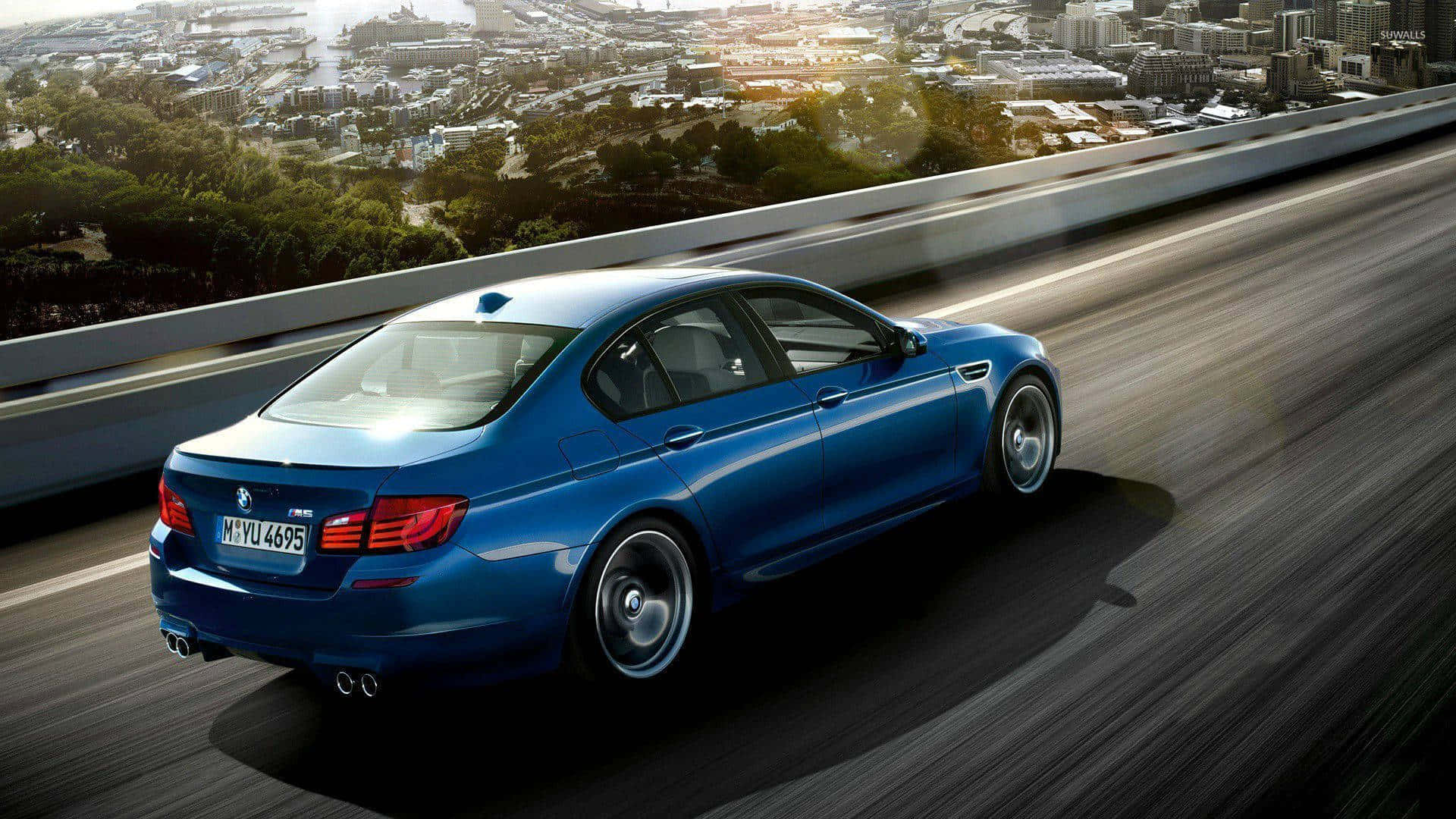 1080p M系列背景：路上的藍色BMW M5 壁紙 — 一輛時尚的藍色BMW M5在高速公路上飛馳，下方是廣闊的城市景觀和綠地，沐浴在金色的陽光下。