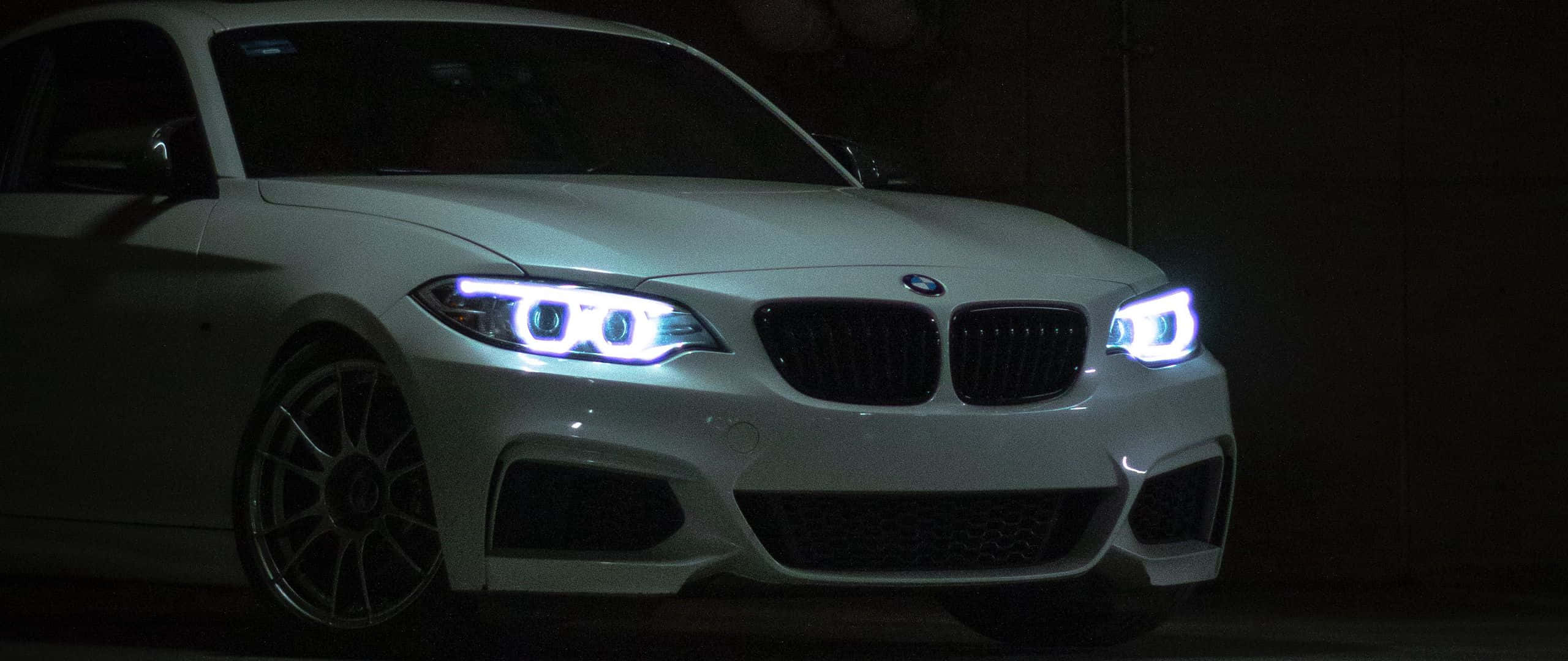 1080p M系列背景：黑夜中的白色BMW 3系 壁紙 — 一輛白色BMW跑車在黑暗的車庫中發出明亮的藍色車頭燈，在其流線型車身上投下銳利的陰影。