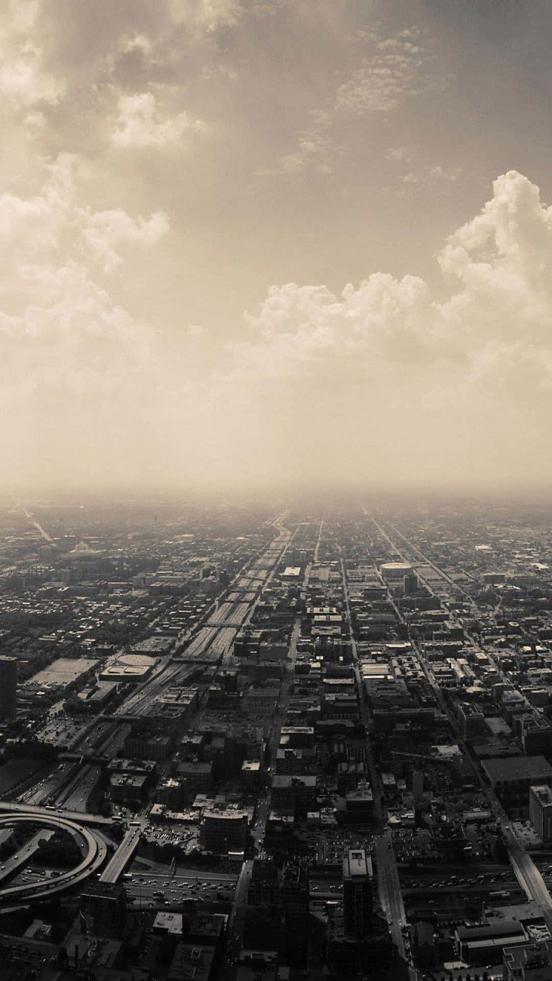 1080x1920 雲層低垂的陰鬱城市 壁紙 — 在戲劇性的多雲天空和薄霧下，廣闊城市網格的懷舊褐色調鳥瞰圖。