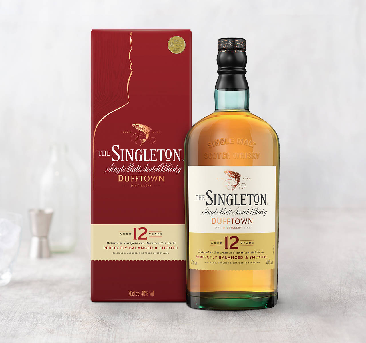 杜夫敦12年陳釀紅蓋威士忌 壁紙 — 一瓶The Singleton 12年單一麥芽蘇格蘭威士忌，旁邊放著它的紅色盒子，在淺色木質表面上，背景有吧檯工具，呈現柔和焦點。