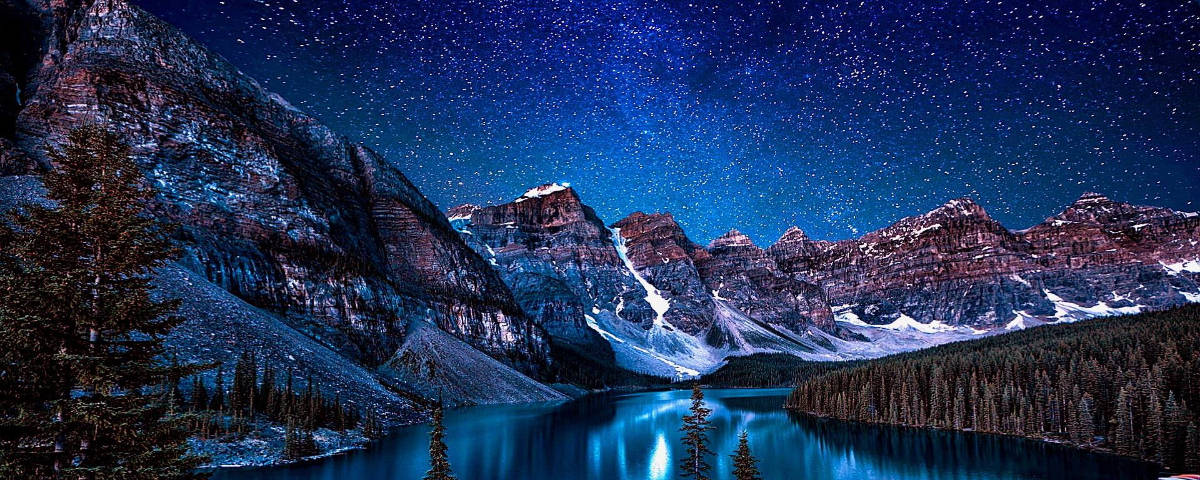 1200x480 星空覆蓋雪山 壁紙 — 繁星點點的夜空倒映在寧靜的山間湖泊中，四周環繞著白雪覆蓋的山峰和常綠樹木。