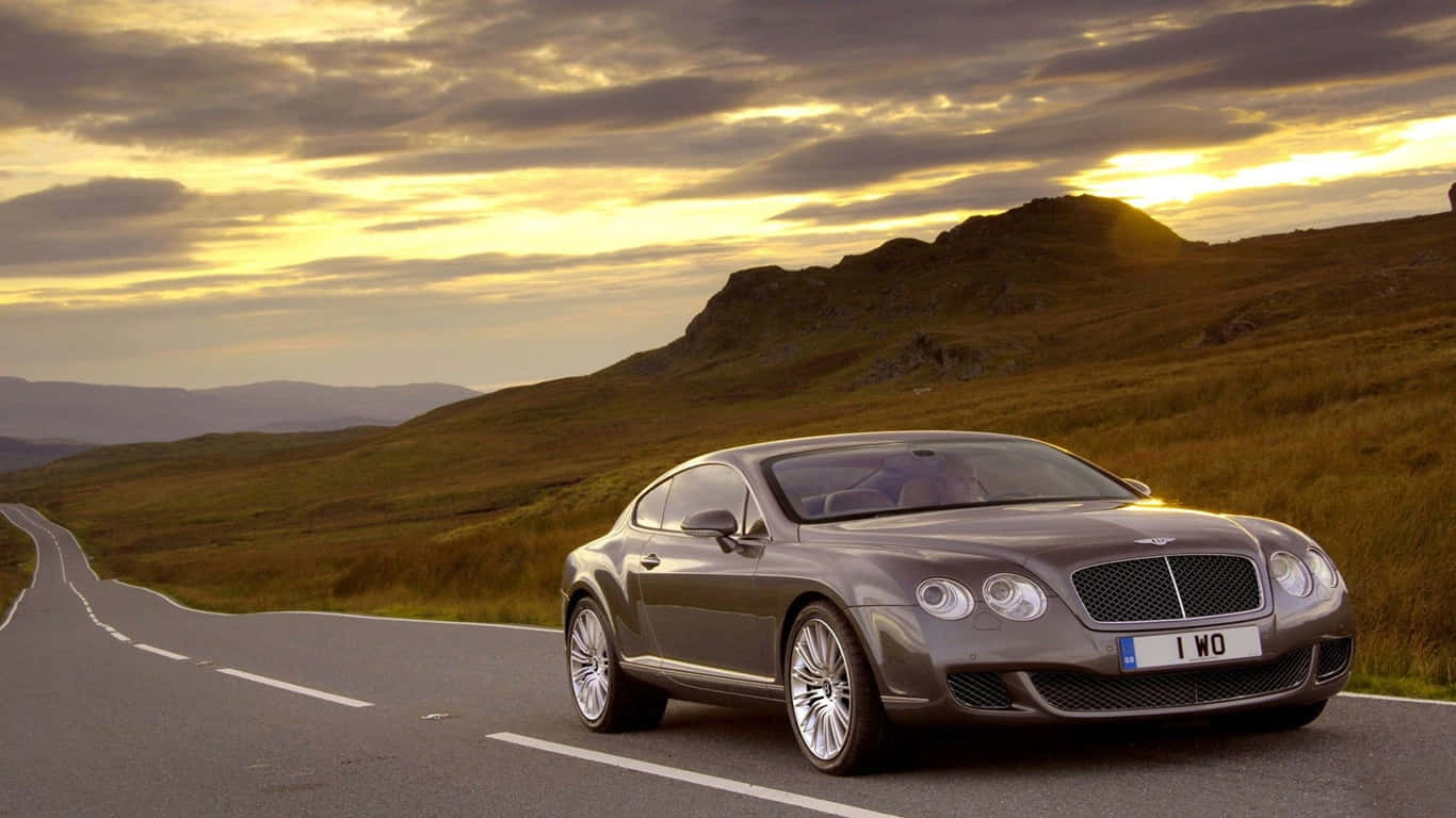 1366x768 2003 賓利 Continental GT 灰色車身背景 壁紙 — 一輛流線型銀色賓利轎跑車沿著蜿蜒的山路行駛，在金色夕陽和戲劇性的雲彩下。