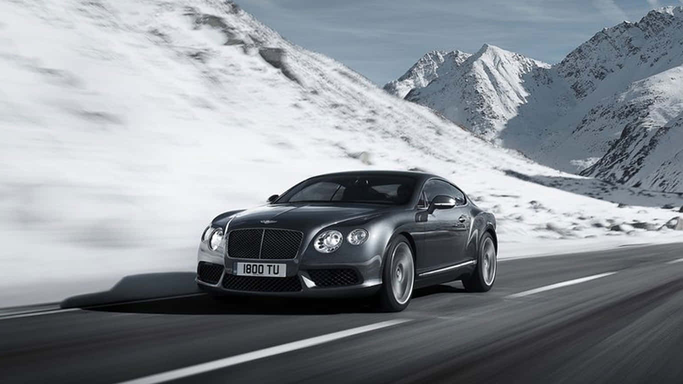 1366x768 2017 年賓利 Continental GT Speed 雪山背景 壁紙 — 一輛時尚的銀色賓利 Continental GT 在晴朗藍天下，沿著蜿蜒的山路疾馳，兩旁是白雪覆蓋的山峰。
