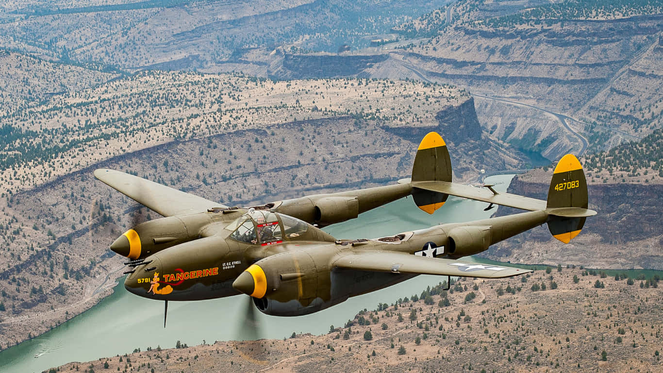 1366x768 巨型客機背景 Lockheed P-38 壁紙 — 一架帶有黃色點綴的復古雙引擎軍用飛機在崎嶇的峽谷景觀上空翱翔，下方有蜿蜒的河流。