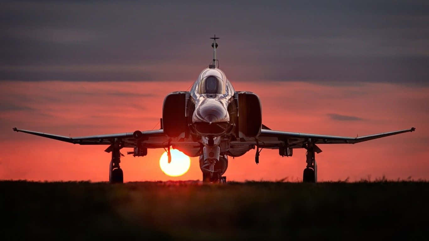 1366x768 巨型客機背景：跑道日落 壁紙 — 一架戰鬥機面向鏡頭，夕陽在其後方落下，在天空中投射出熾熱的橙色光芒。