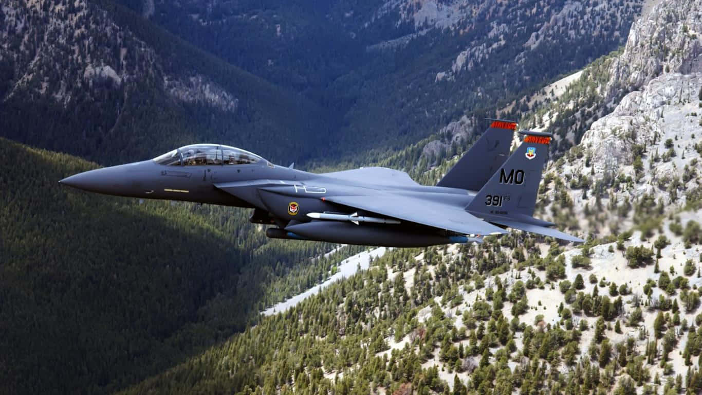 1366x768 巨型客機背景 綠色山巒 壁紙 — 一架灰色戰鬥機在明亮的日光下，低空飛過森林覆蓋、山頂積雪的山脈。