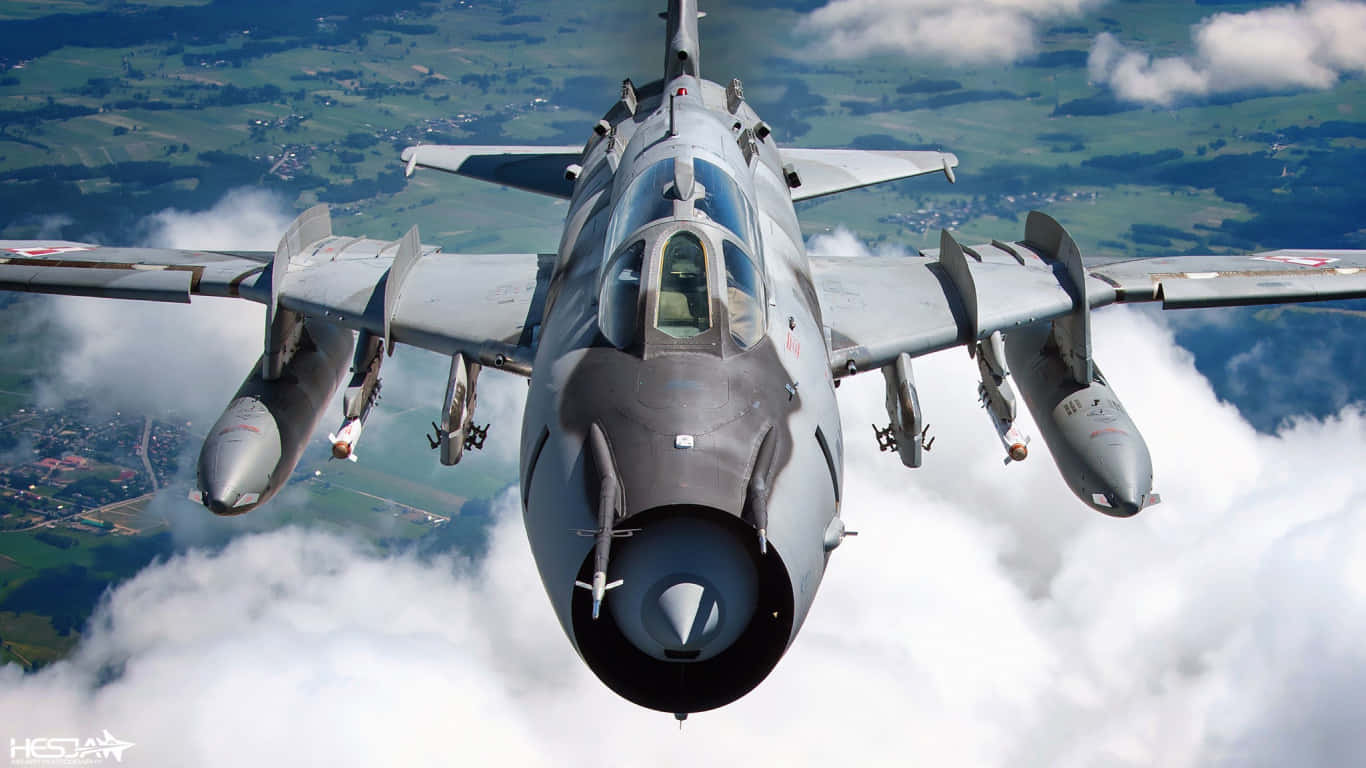 1366x768 巨型客機背景：蘇霍伊 Su-17 壁紙 — 一架灰色戰鬥機在蓬鬆的白雲上方翱翔，下方是綠色的鄉村。