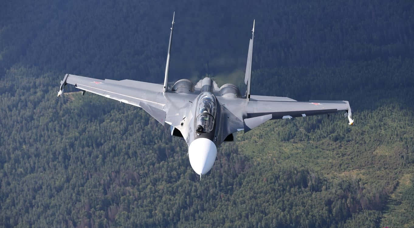 1366x768 巨型客機背景：森林地面 壁紙 — 一架灰色戰鬥機在晴朗天空下飛越森林覆蓋的山脈。