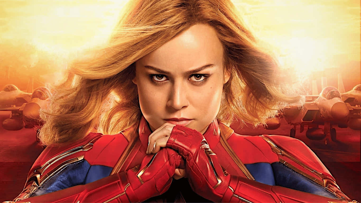 1366x768 捕捉漫威《卡蘿·丹弗斯》握拳背景 壁紙 — Captain Marvel 自信地站立，身穿紅藍戰衣，背後有戰鬥機，襯托著發光的日落天空。