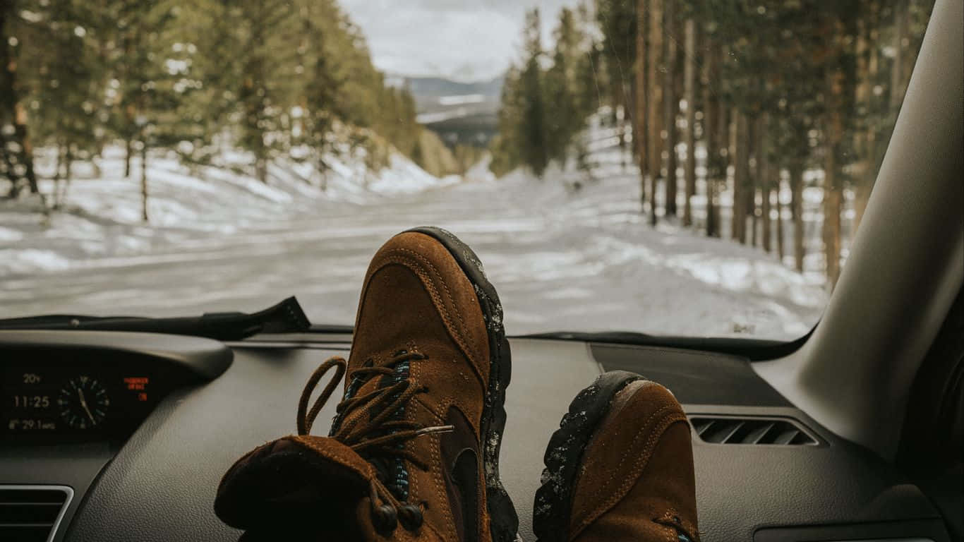 棕色登山靴擱在汽車儀表板上，面向一條積雪的森林道路。