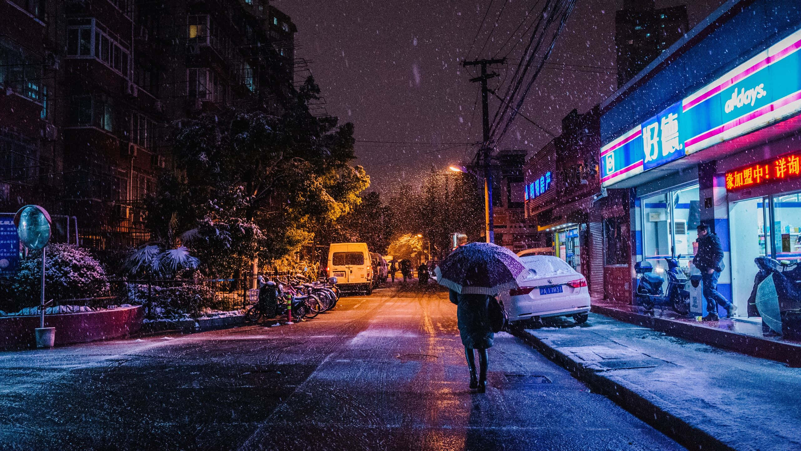 1440p 雪夜獨行街景背景 壁紙 — 一個孤獨的身影在夜晚的雪城市街道上行走，在發光的霓虹燈招牌和街燈下撐著雨傘。