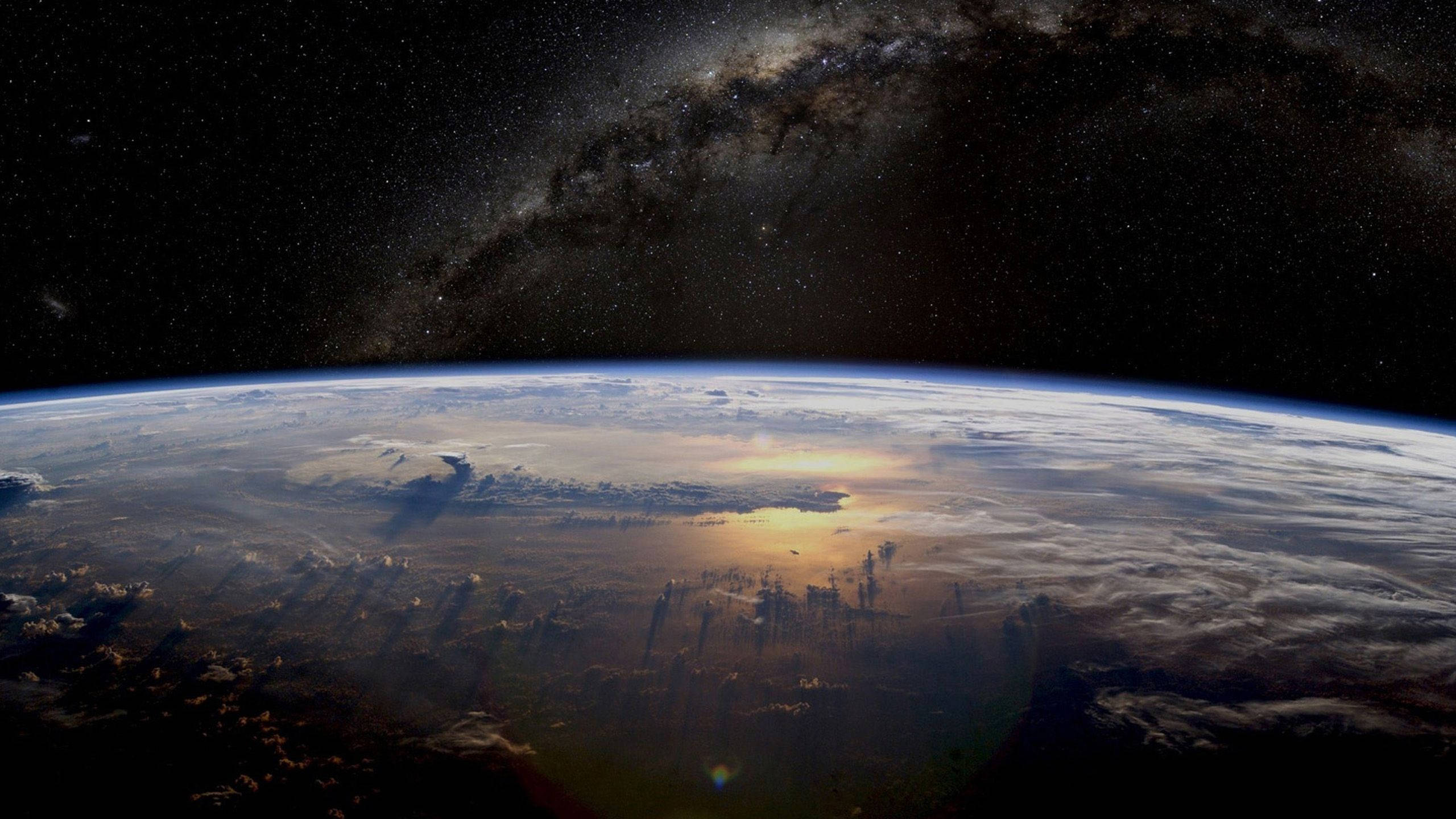 1440p 高畫質太空中的星球 壁紙 — 地球在星空下彎曲，銀河拱起，陽光在雲層上閃爍。