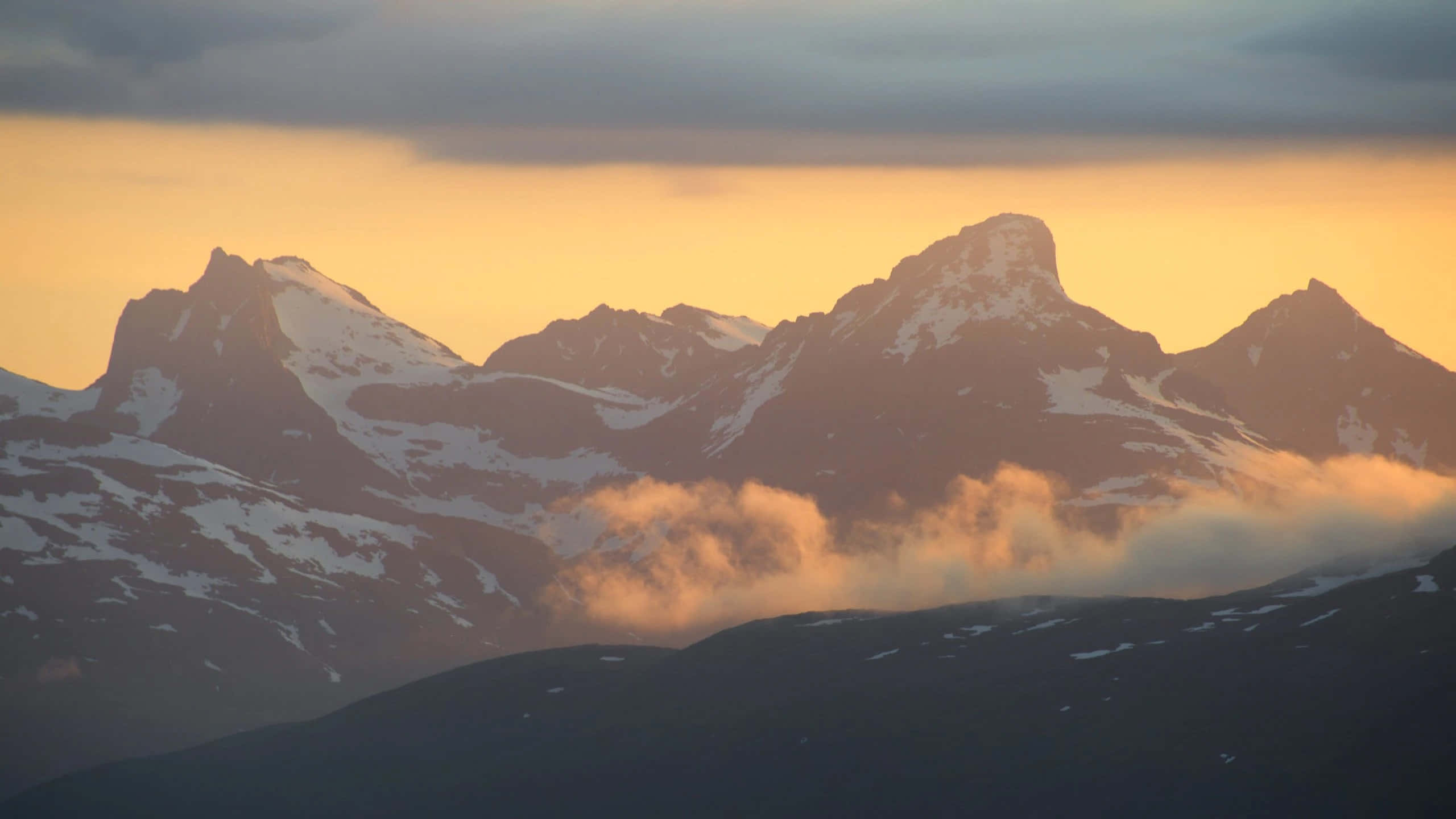 1440p 旅行 Tromsø 背景 壁紙 — 積雪的山峰在日落時分聳立於發光的雲層之上，溫暖的金色光芒照亮了天空。