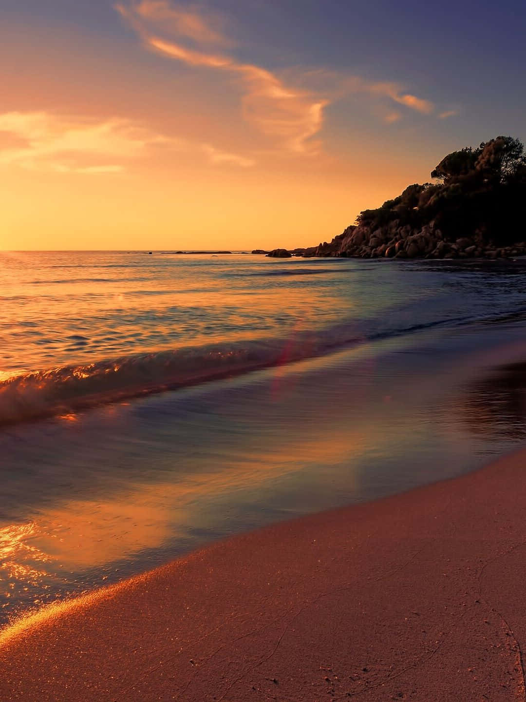 1440p 自然背景：海灘夕照 壁紙 — 金色夕陽海浪輕輕拍打著沙灘，旁邊是點綴著樹木的岩石懸崖。