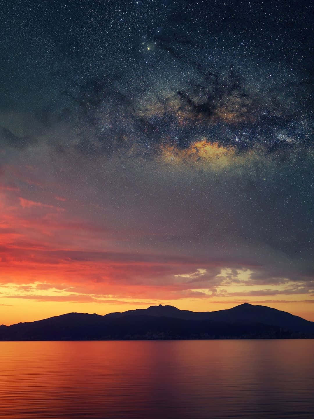 1440p 自然背景：星際天空與山巒 壁紙 — 令人嘆為觀止的暮色天空，帶有鮮豔的橙色雲彩和閃爍的銀河，拱形跨越剪影山脈和寧靜的水面。