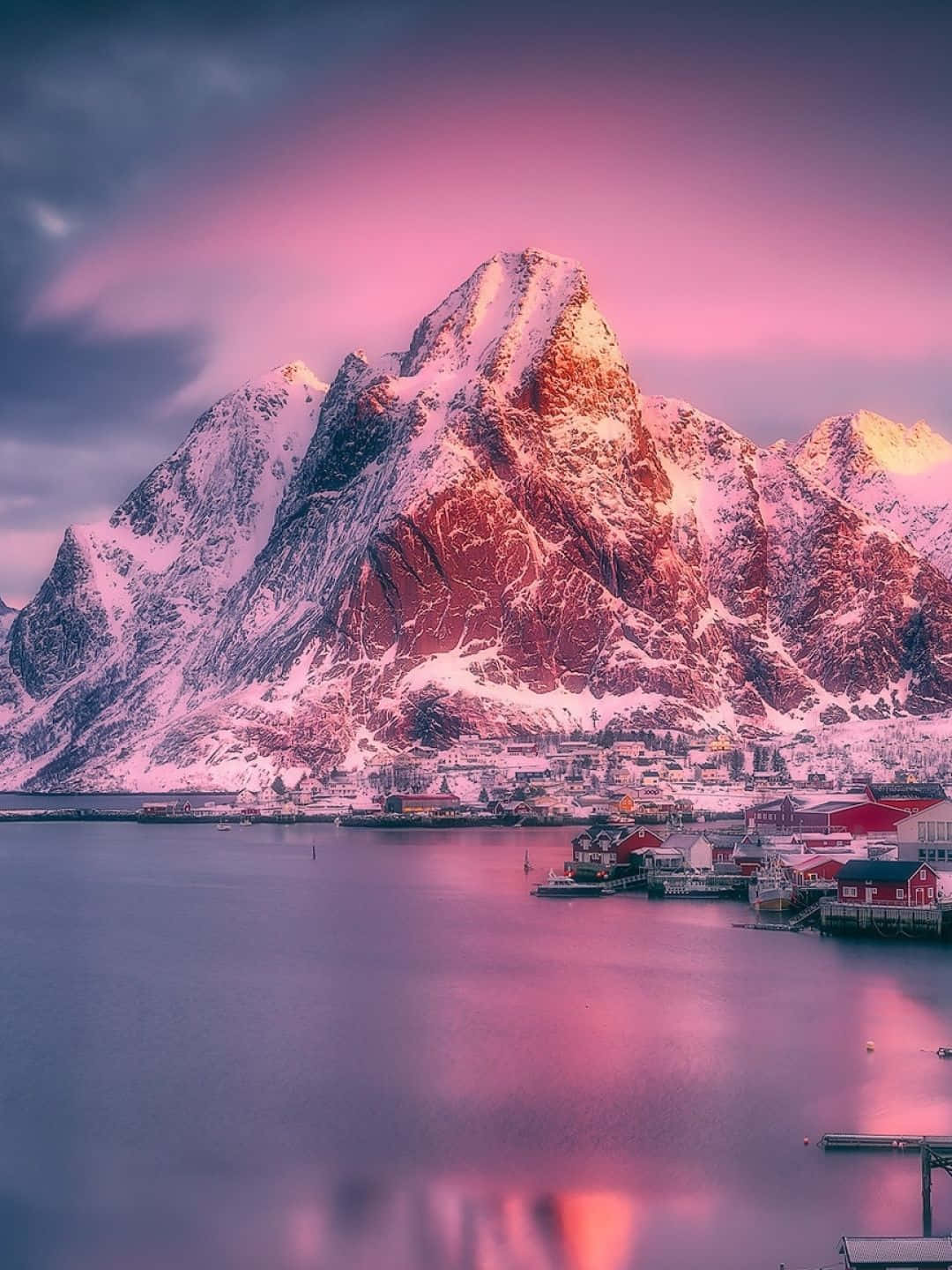 1440p 自然背景：羅弗敦群島雪峰與粉紅雲霞 壁紙 — 積雪的山峰在日落時分泛著粉紅色的光芒，映照在如鏡面般的峽灣上的一個寧靜漁村。