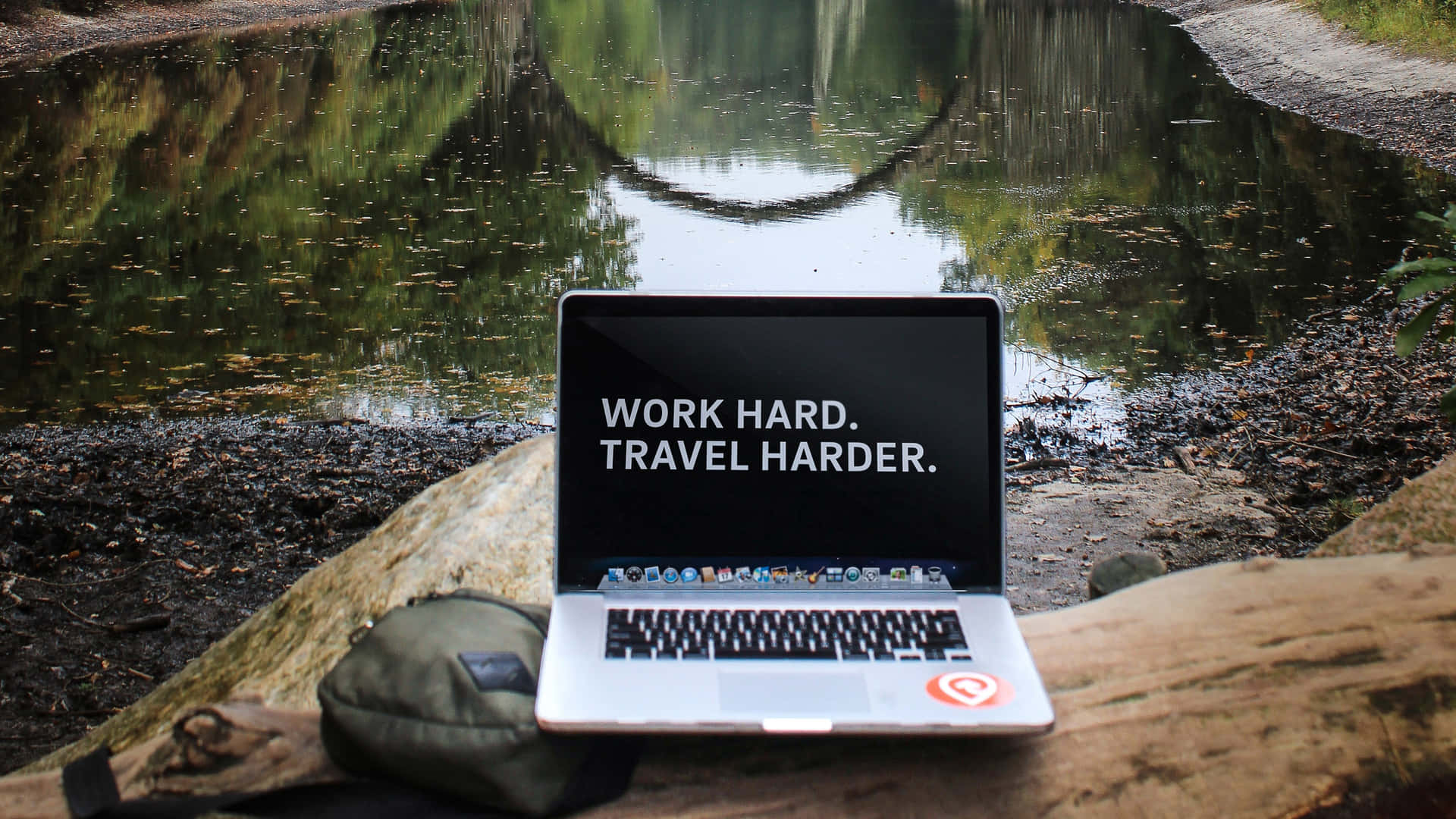 池塘旁圓木上的手提電腦，附有勵志語句 work hard travel harder