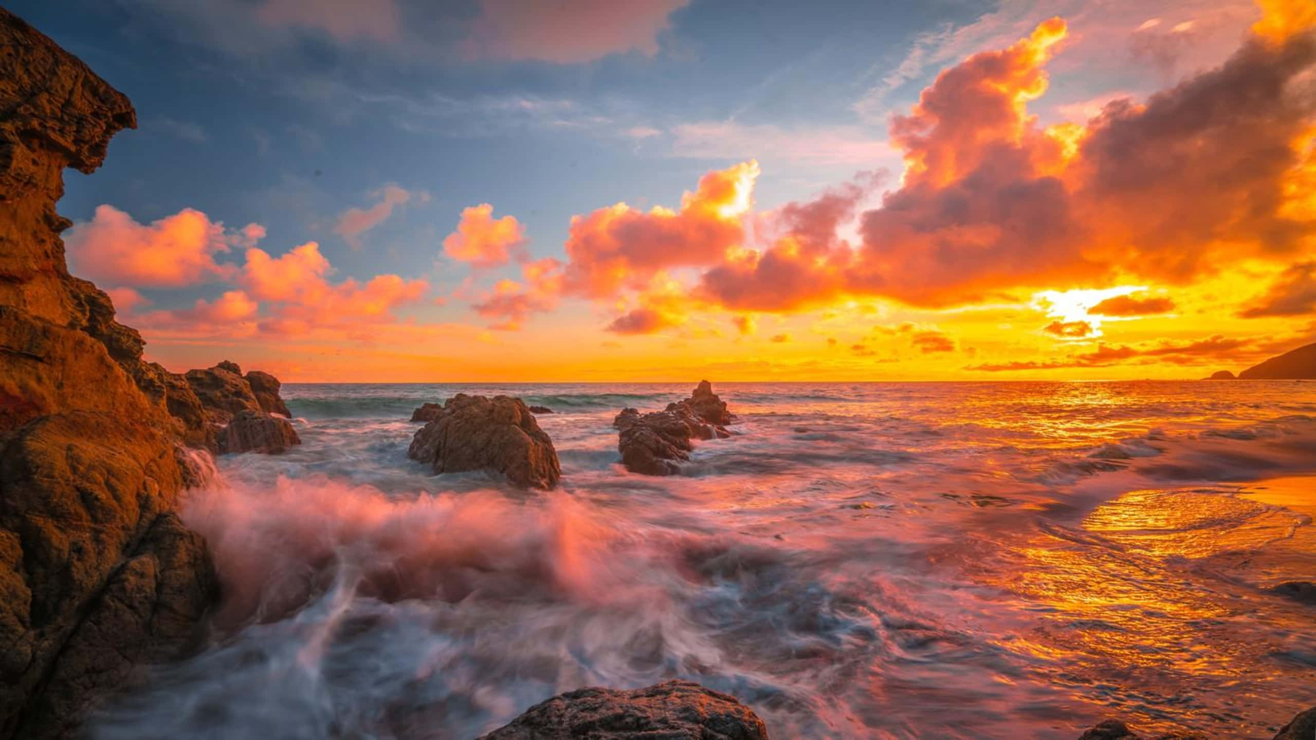 1440p 馬利布日落海浪背景 壁紙 — 夕陽將天空染成火熱的橙色和粉紅色，海浪拍打著崎嶇的海岸岩石。