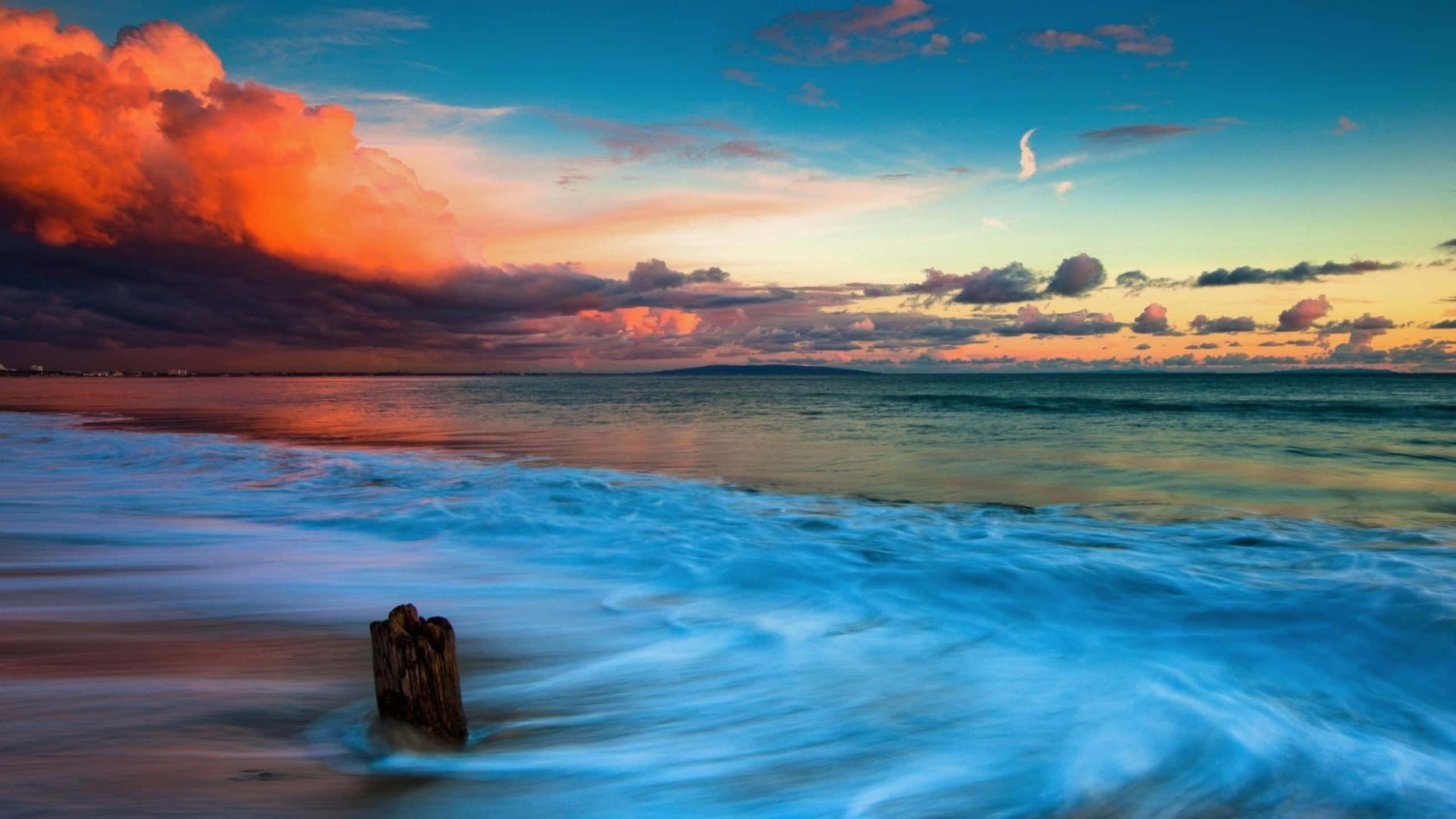 1440p 馬利布退潮海浪背景 壁紙 — 一根孤獨的風化木樁矗立在泡沫翻騰的海浪中，背景是戲劇性的日落天空，有火紅的橙色雲朵和一彎新月。