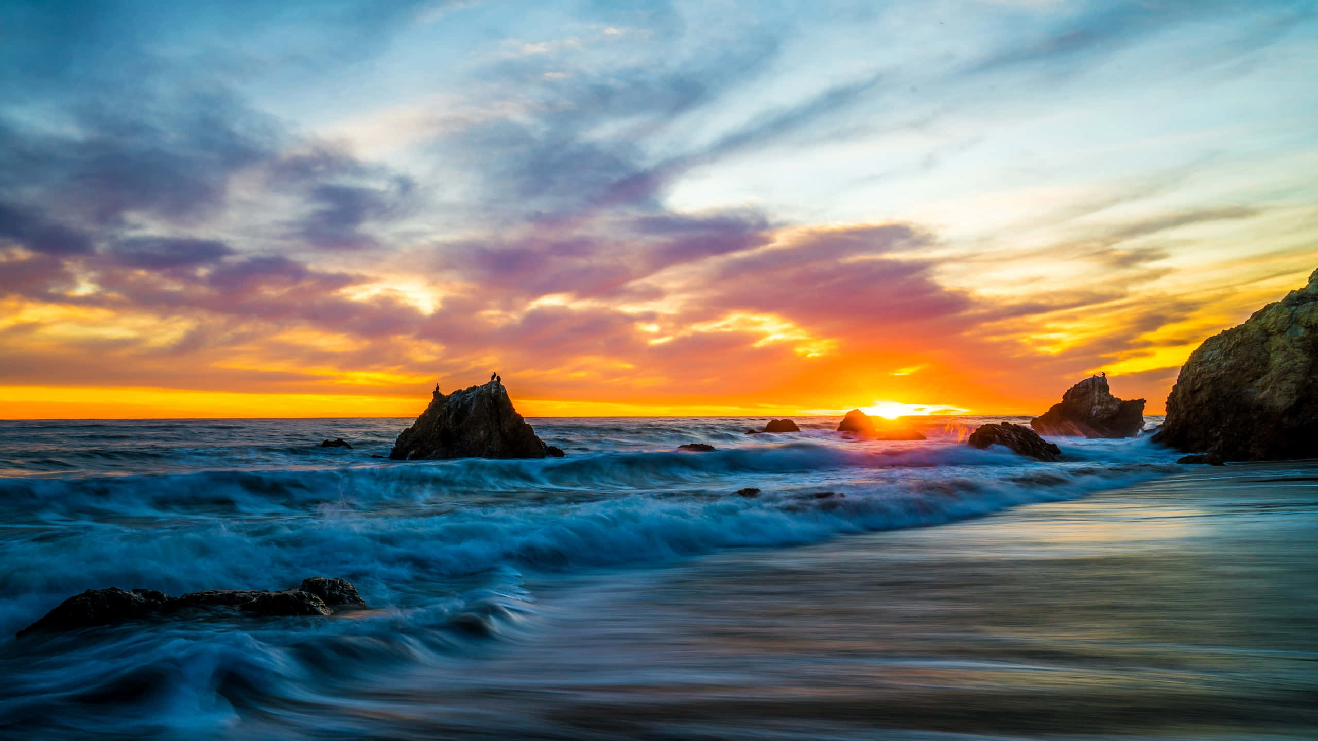 1440p 馬里布海灘浪花背景 壁紙 — 夕陽將天空染成火熱的橙色和紫色，海浪拍打著崎嶇的海岸岩石。