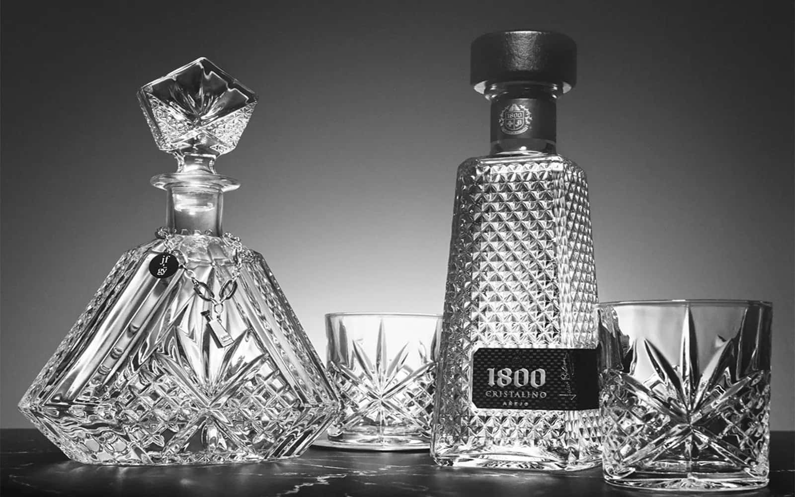 1800 蒂克利亞水晶陳年龍舌蘭酒配水晶醒酒器 壁紙 — 優雅的黑白靜物，展示標有1800 Cristalino的水晶醒酒瓶及配套酒杯，置於深色大理石檯面上。