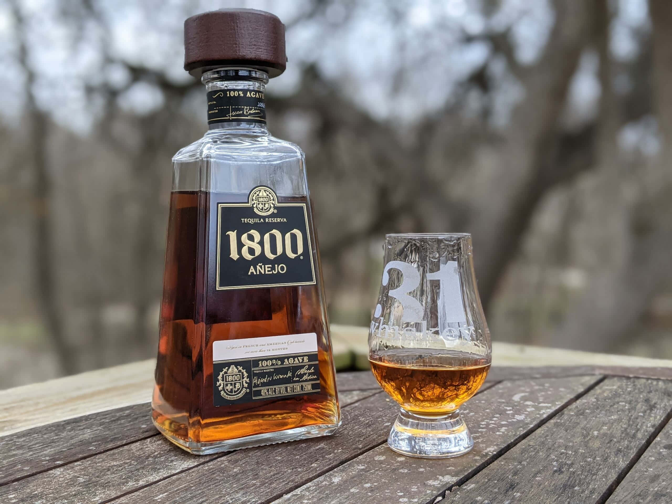 1800 蒂奎拉陳年款酒瓶與玻璃杯 壁紙 — 一瓶1800 Añejo龍舌蘭酒和一個品酒杯放在戶外一張風化的木桌上，背景是模糊的樹木。