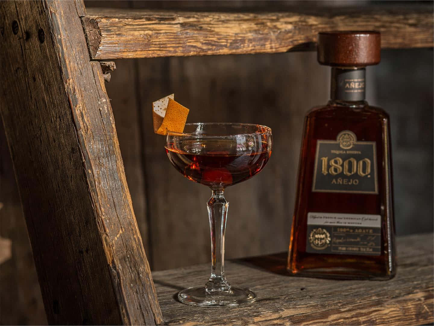 墨西哥品牌1800龍舌蘭酒Reserva Anejo與木質桌面玻璃杯 壁紙 — 一杯豐富的琥珀色雞尾酒盛載於笛型杯，飾以橙皮，旁邊放著一瓶1800 Arejo龍舌蘭酒，置於質樸的木質表面上。
