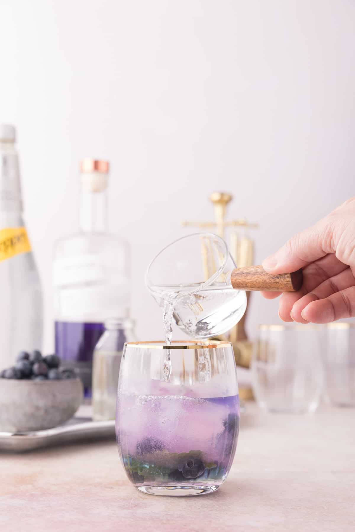 帝后1908金酒搭配蘇打水 壁紙 — 一隻手將清澈液體倒入一個帶有紫色藍莓飲料和金色邊緣的玻璃杯中，置於柔軟的粉紅色表面上。