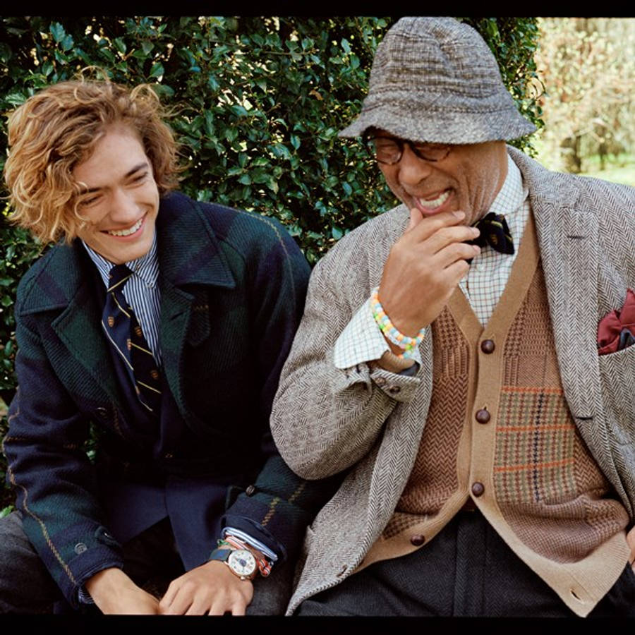 1920年代風格 Ralph Lauren 壁紙 — 兩個穿著時尚層次服裝的男人在茂密的綠色樹籬背景下分享歡樂的笑聲。