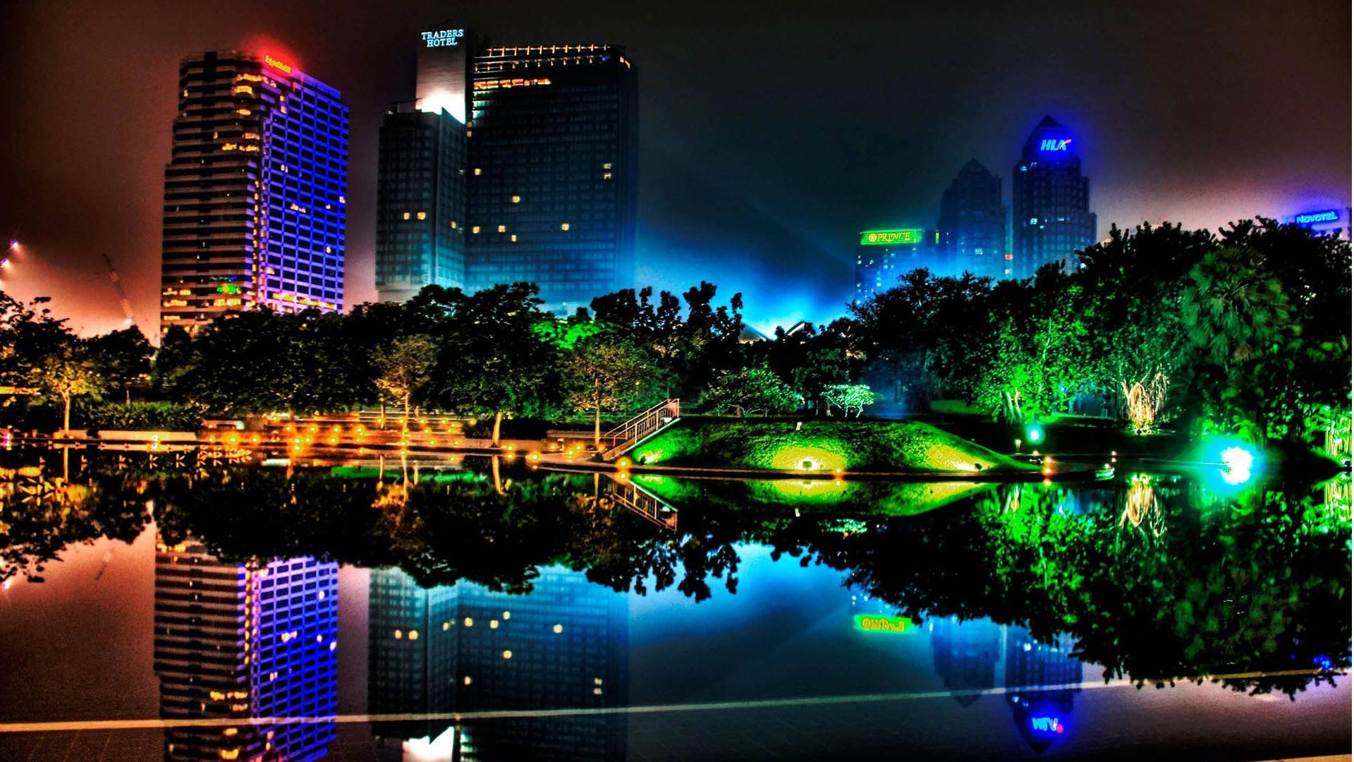 1920 X 1080 夜間城市霓虹燈摩天大樓 壁紙 — 充滿活力的夜間城市景觀，發光的摩天大樓倒映在平靜的湖面上，周圍環繞著發光的綠色樹木。