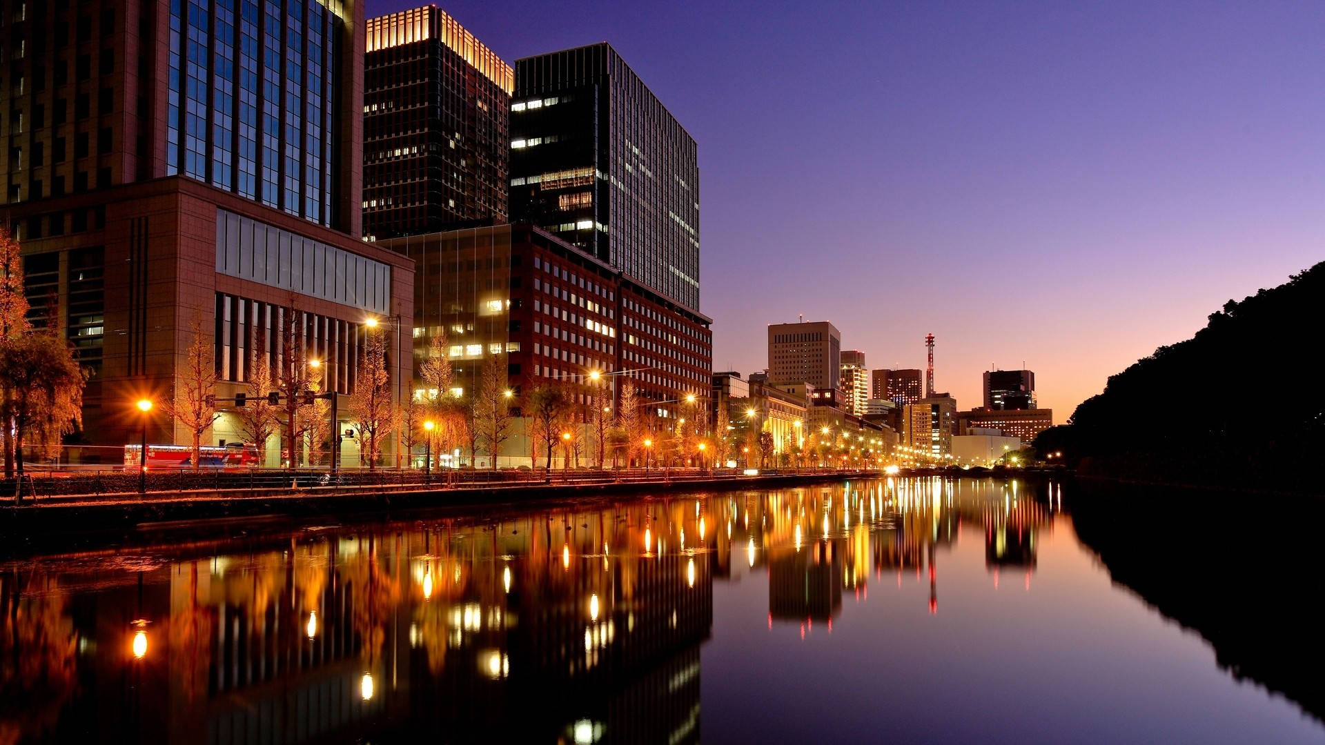1920 X 1080 夜間東京城市 壁紙 — 暮色中的城市天際線，溫暖的路燈和建築物倒影在平靜的水面上閃爍。