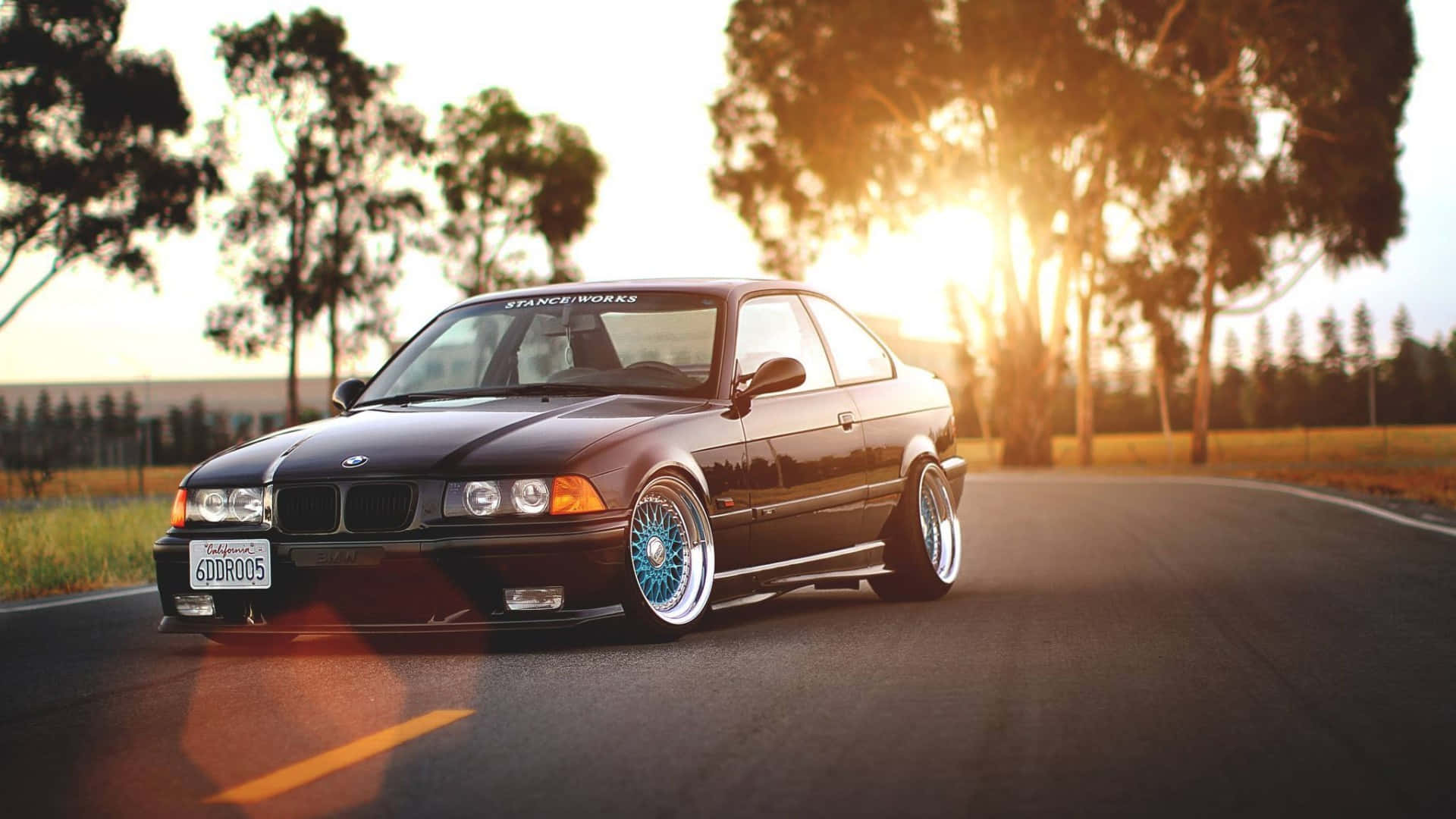 金色日落時分，一輛配藍色輪圈的黑色BMW E36雙門轎跑車停泊在道路上。