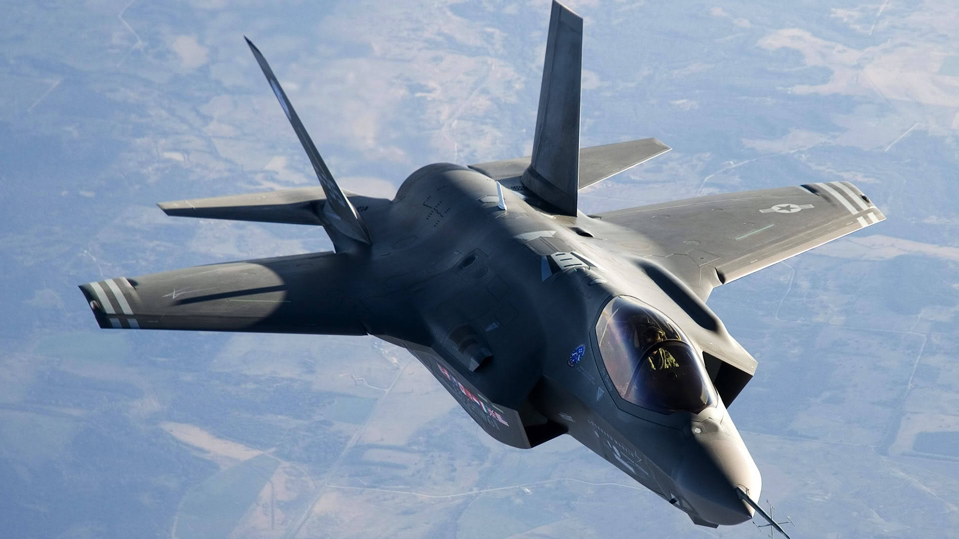 1920x1080 F-35A 隱形戰鬥機 壁紙 — 一架流線型深灰色戰鬥機在晴朗藍天下翱翔於拼布般的地景之上。
