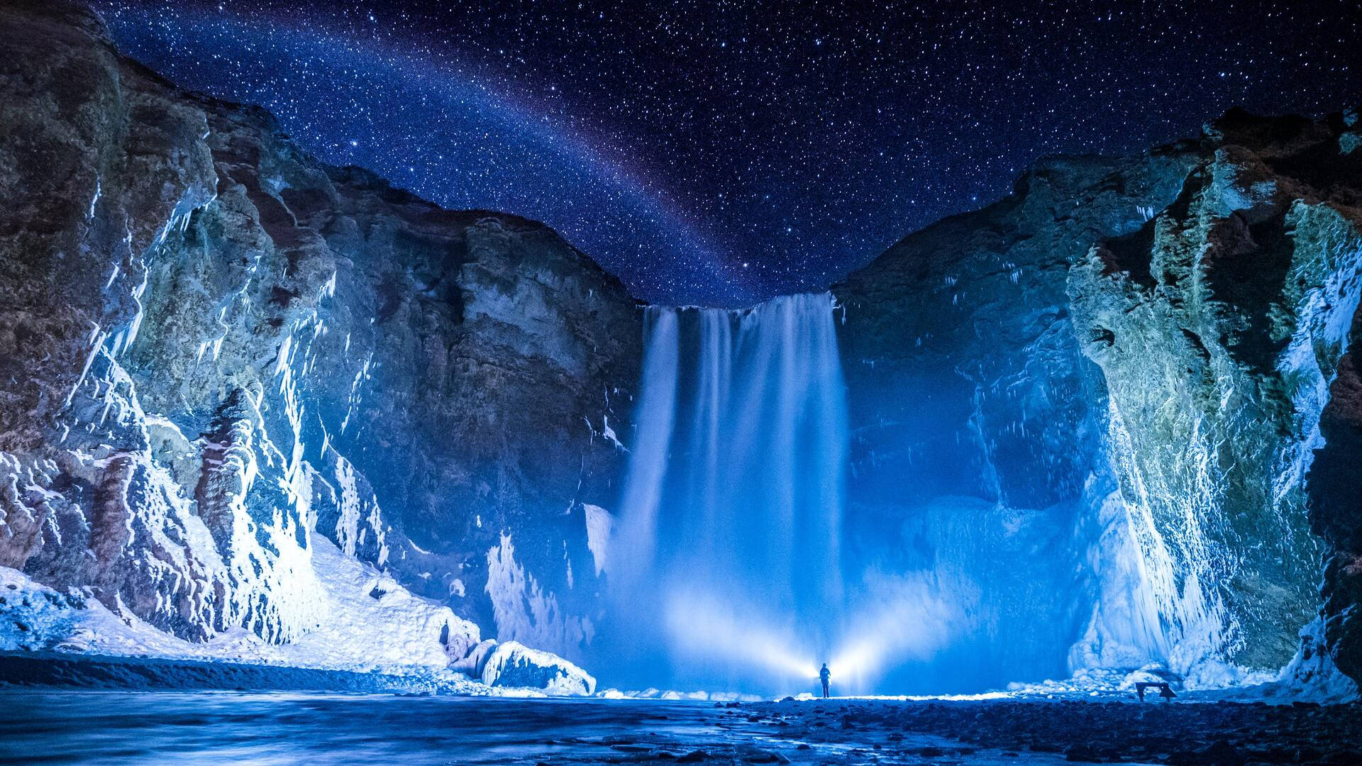 1920x1080 HD 斯科加瀑布夜景 壁紙 — 在冰凍的峽谷中，一個孤獨的身影站在星空下發光的瀑布下。