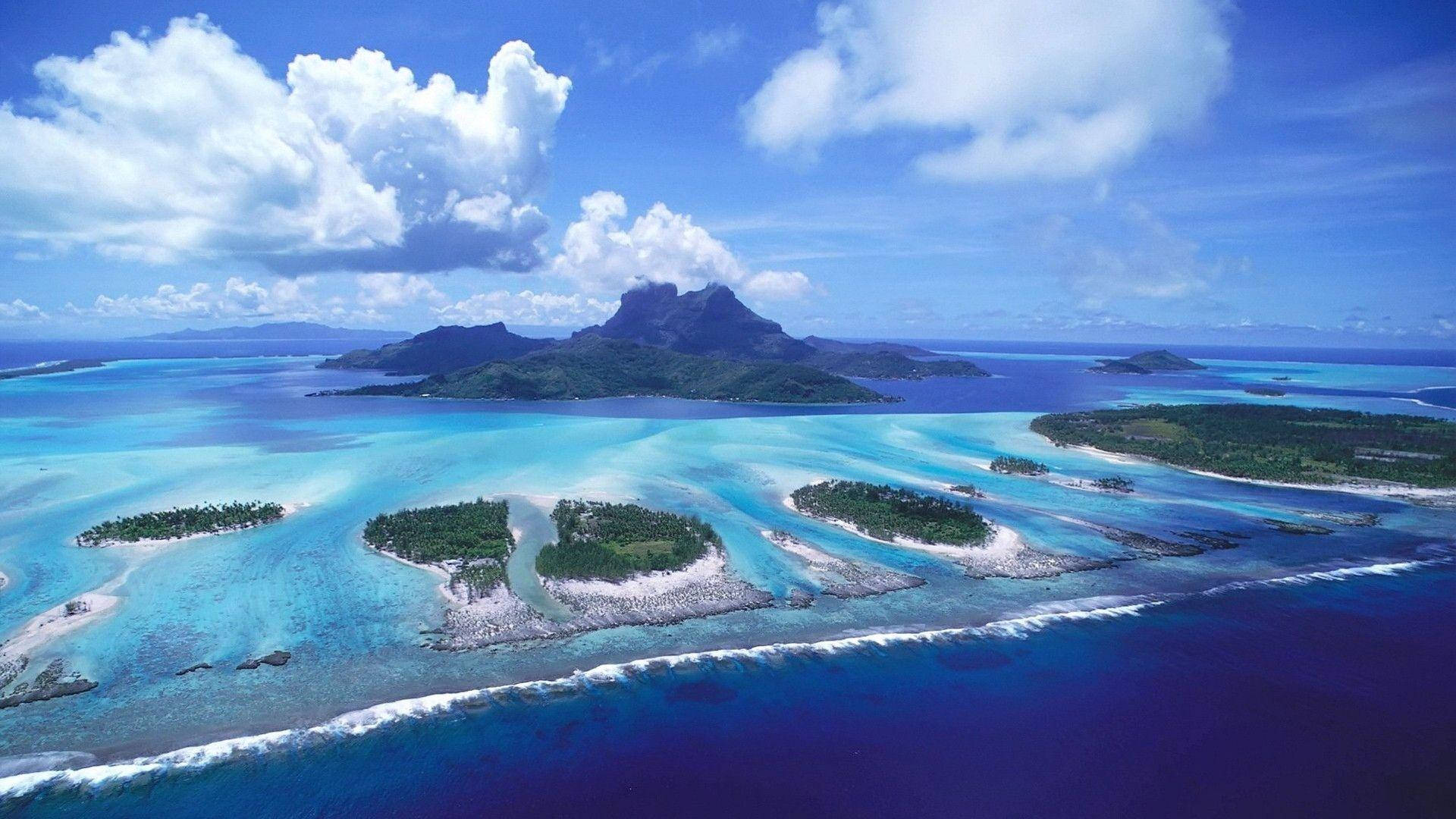 1920x1080 全高清海島鳥瞰 壁紙 — 在明亮的藍天和蓬鬆的白雲下，俯瞰碧綠色的潟湖和鬱鬱蔥蔥的綠色島嶼。