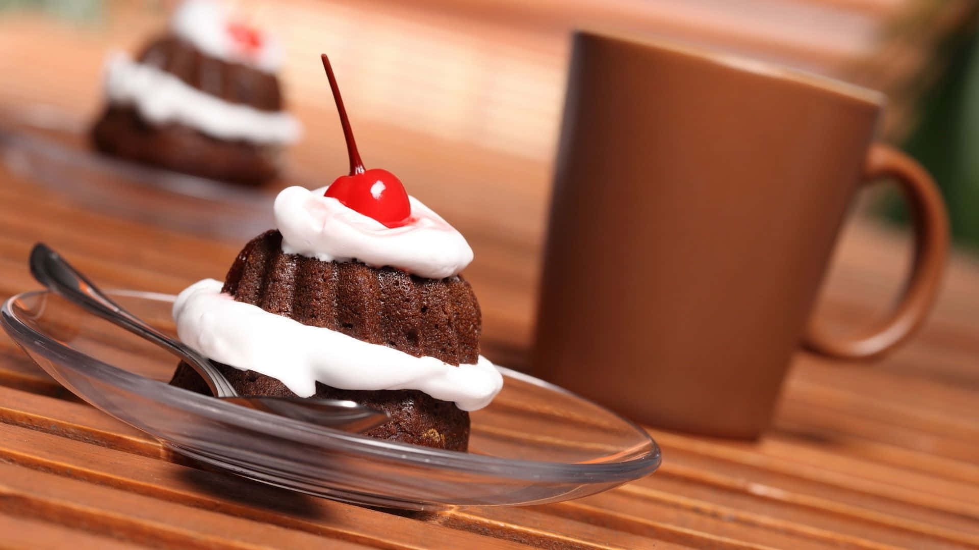 1920x1080 蛋糕背景：迷你 Bundt 蛋糕 壁紙 — 一個豐富的朱古力蛋糕配白色忌廉和櫻桃，放在玻璃碟上，旁邊有一個棕色馬克杯在木桌上。