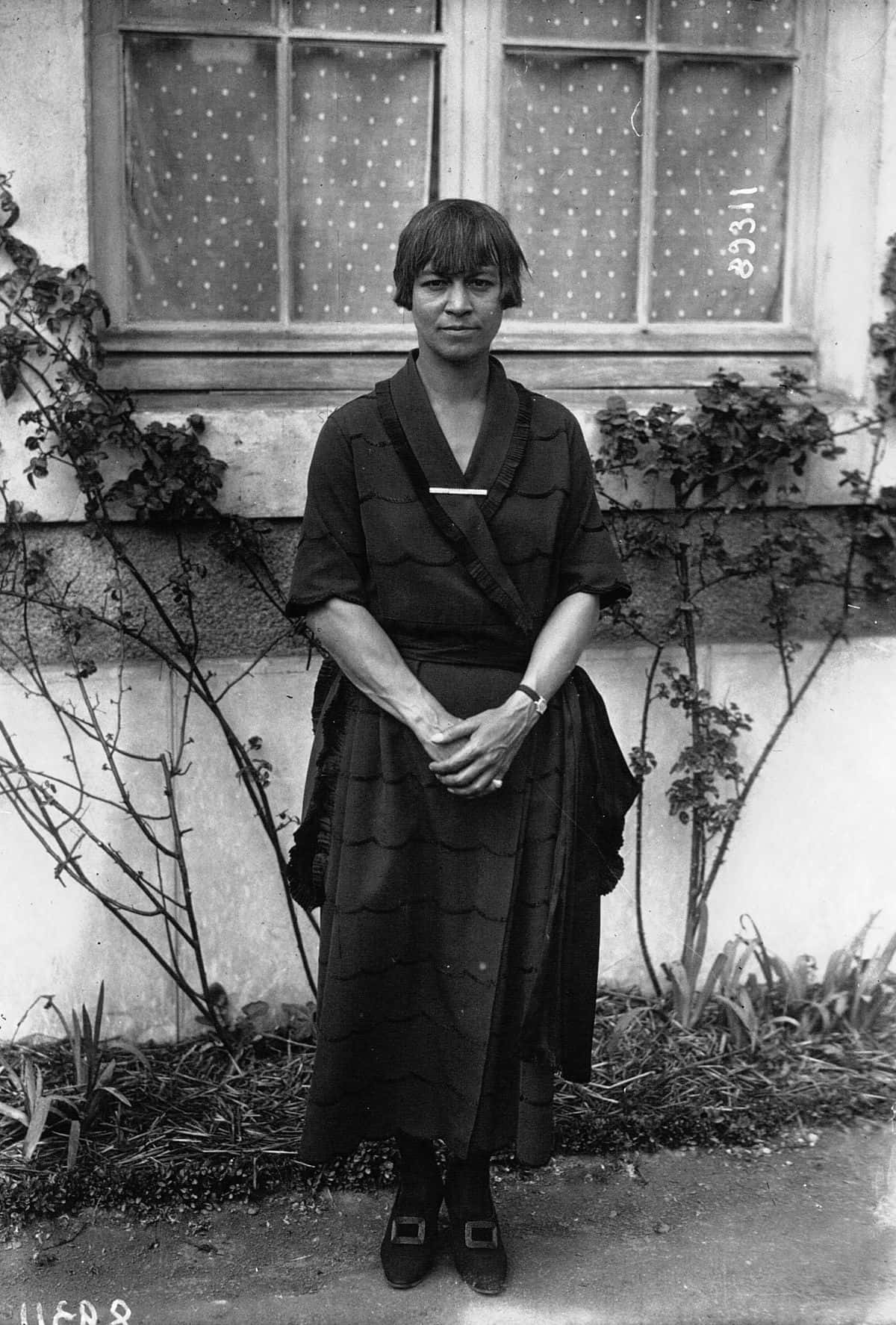 莫拉·馬洛里，約1921年 壁紙 — 一位身穿深色圖案連衣裙的女性平靜地站在帶有攀緣藤蔓和圓點窗簾的窗牆前。