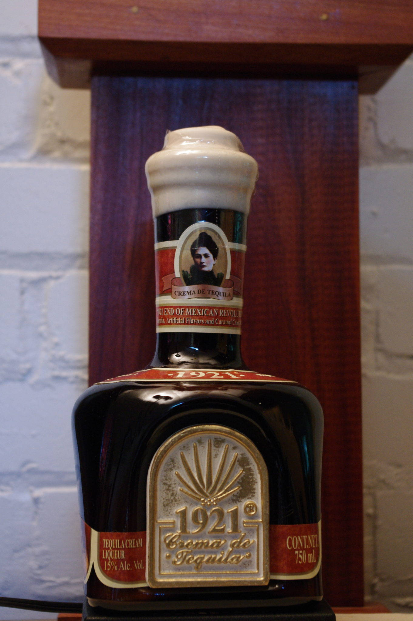 1921奶油龍舌蘭迷你瓶 壁紙 — 一瓶深色玻璃瓶裝的1921 Crema de Tequila，配有奶油色瓶蓋，放在白色磚牆前的木架上。