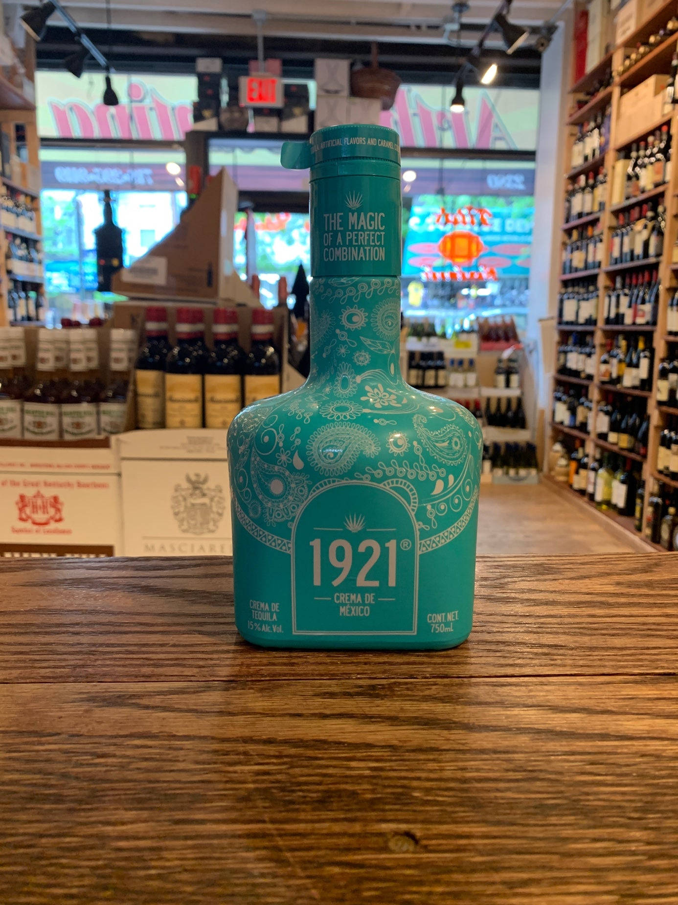1921 龍舌蘭藍瑪雅奶油瓶於酒類商店 壁紙 — 一瓶1921 Crema de Mexico青綠色龍舌蘭酒，飾有華麗的白色圖案，放在溫暖燈光的酒舖的質樸木櫃檯中央。