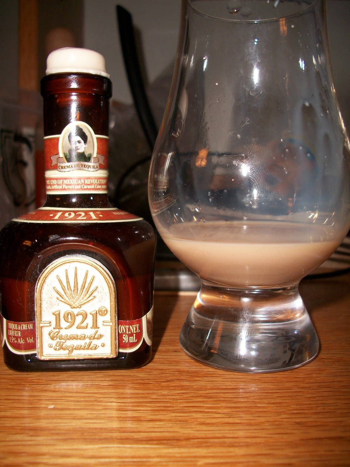 一瓶1921 Tequila Cream Liqueur旁邊有一個玻璃杯，裏面裝著其奶油飲品，放在木上。