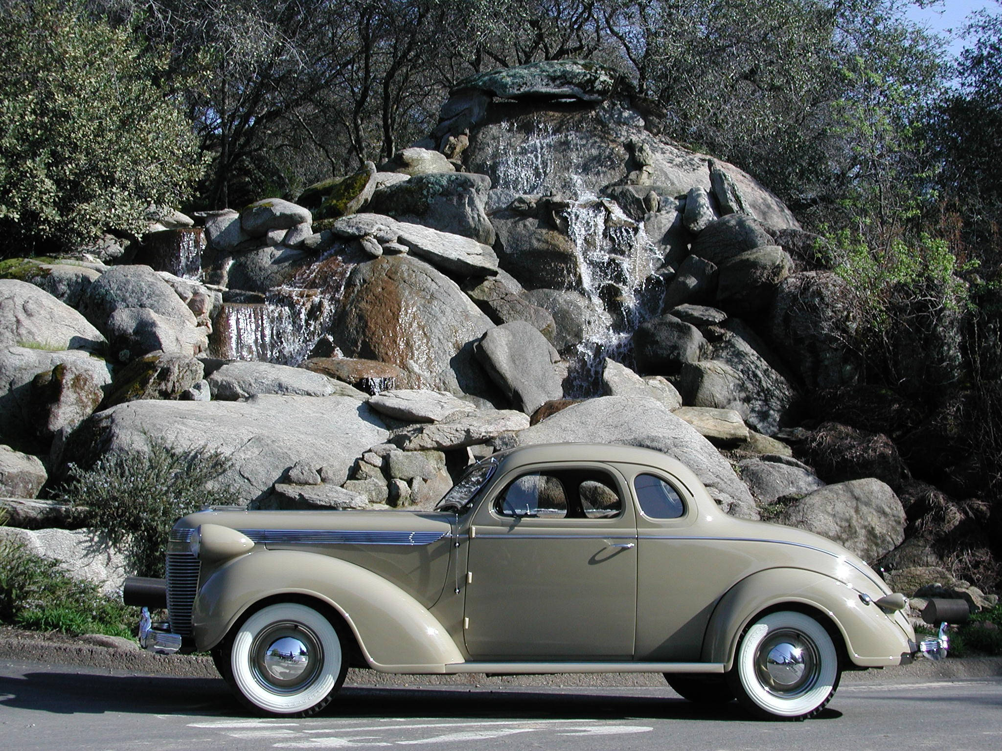 1937年雪佛蘭帝國轎車 壁紙 — 一輛復古米色雙門轎車，帶白色側壁，停泊在陽光穿過樹林、岩石瀑布前。