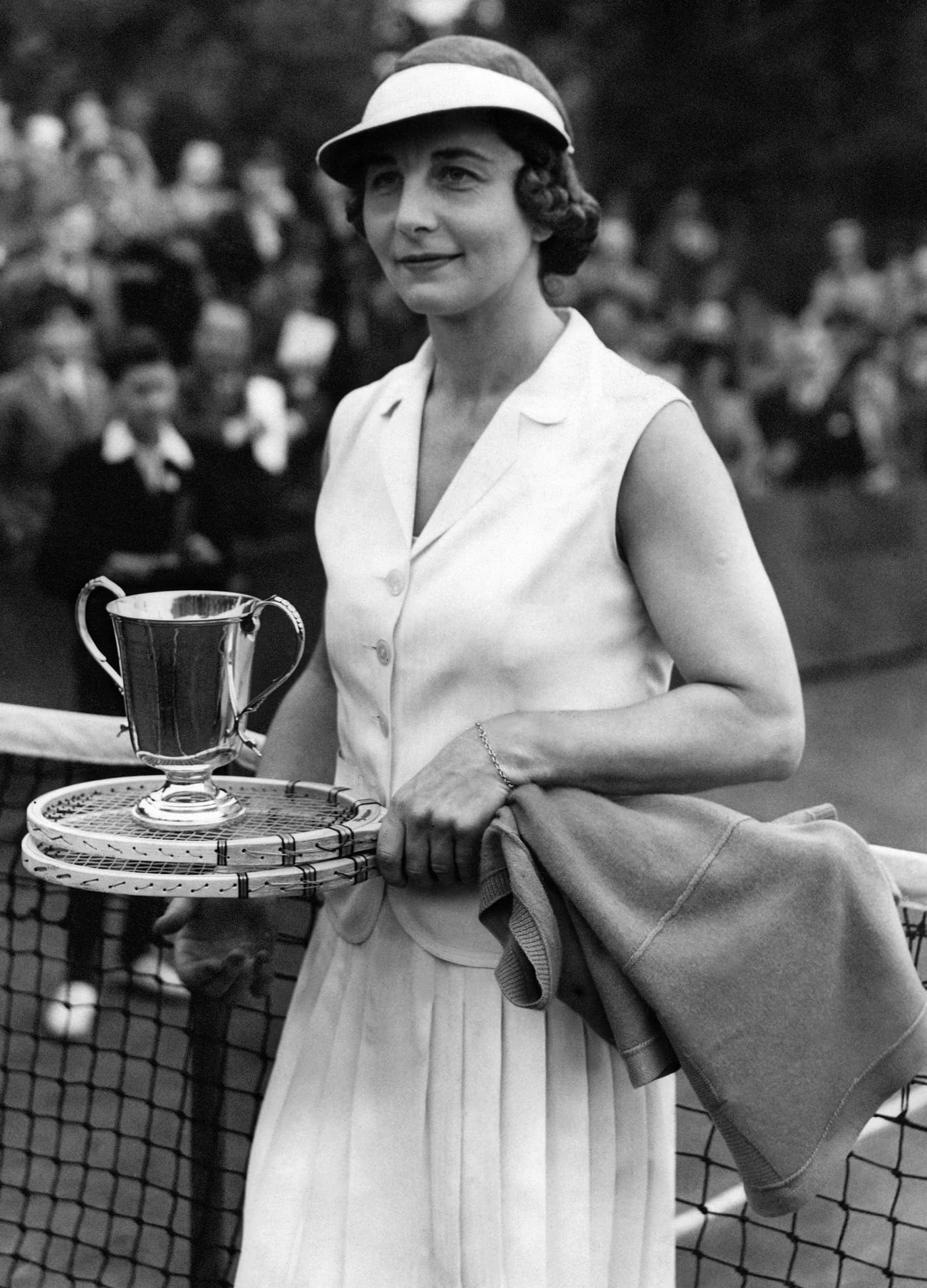 美國網球選手海倫·威爾斯 1938年肖像 壁紙 — 一張復古黑白肖像，一位微笑的網球冠軍在球場上舉起獎盃，身後有觀眾。