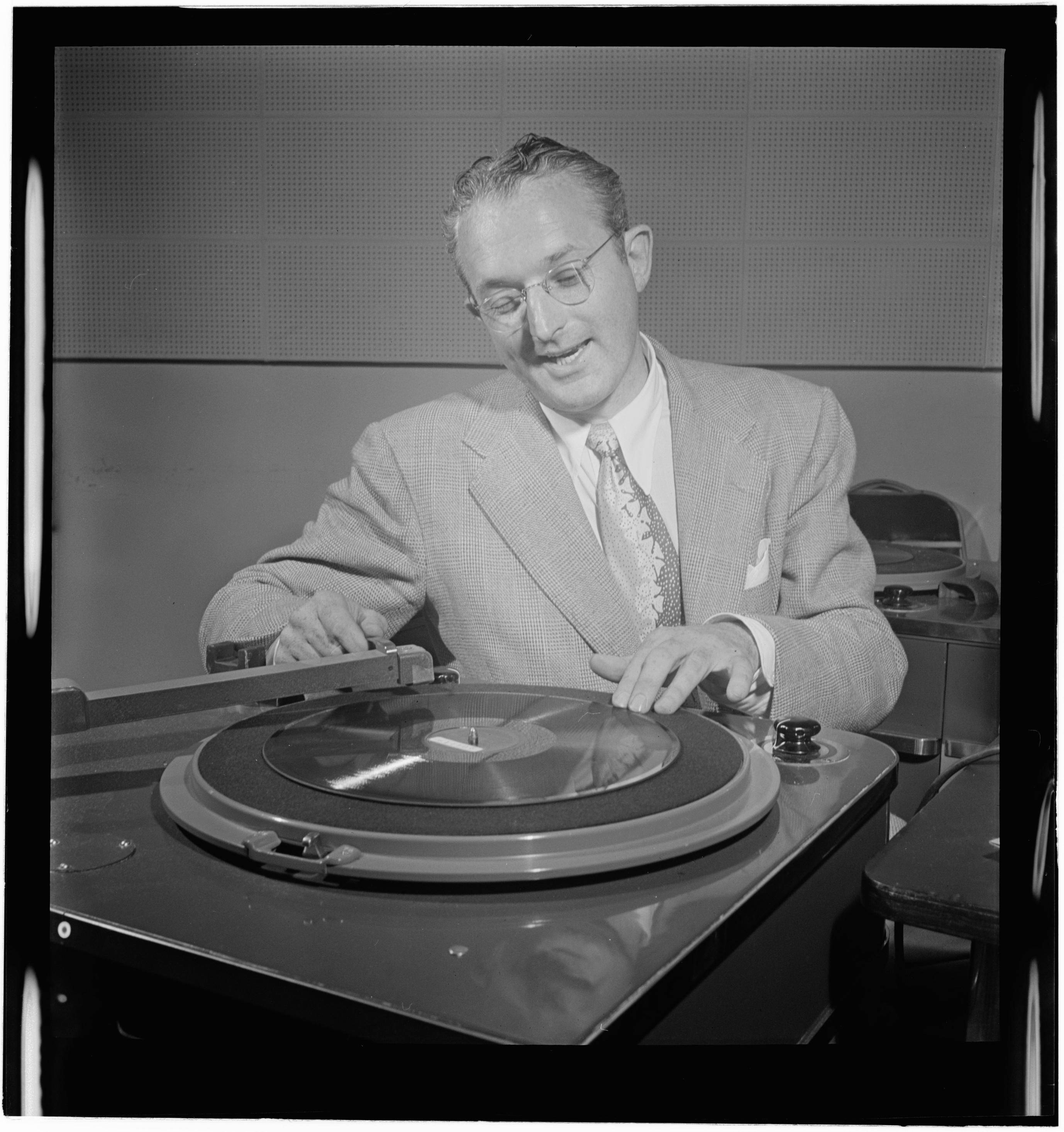 1947年湯米·多西操作留聲機黑白攝影 壁紙 — 一位微笑的西裝眼鏡男在錄音室中小心地將黑膠唱片放在復古唱盤上。