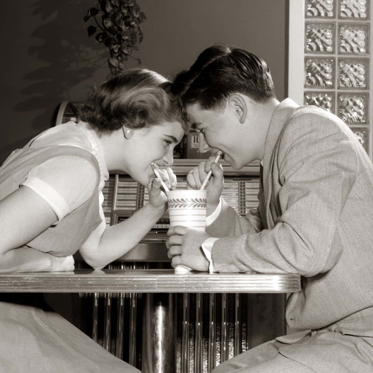 1950年代經典汽車停泊於海灘旁 壁紙 — 一張復古黑白照片，一對年輕情侶在餐廳卡座用吸管分享飲料。