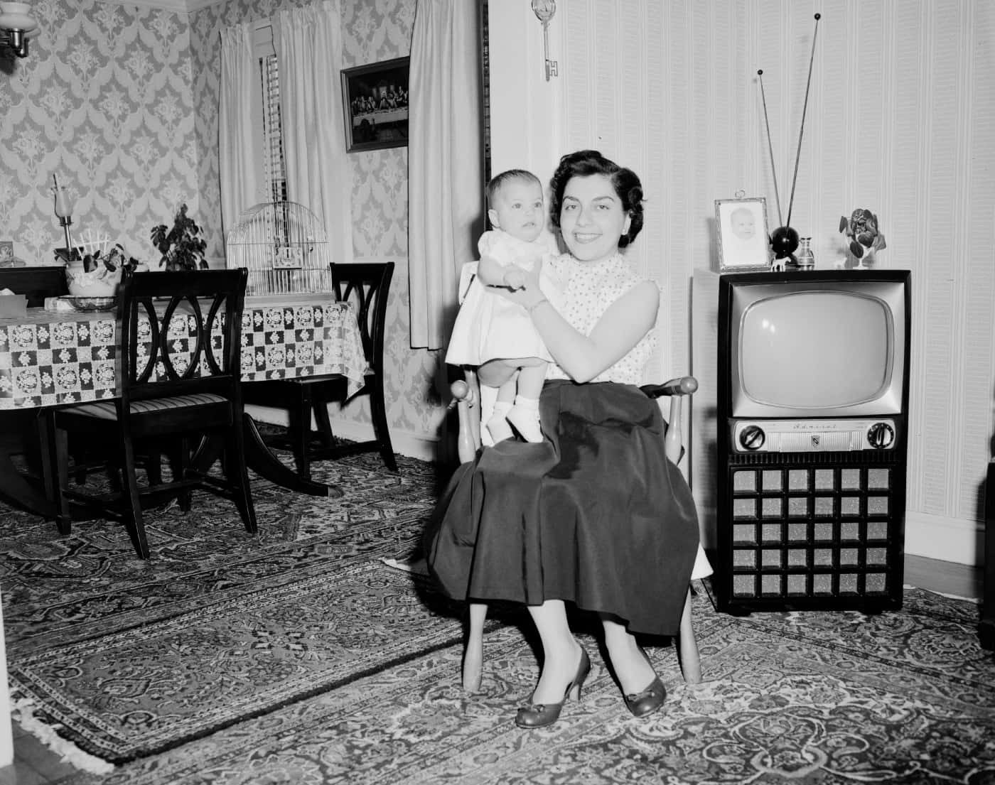 女士在1950年代客廳中抱著嬰兒，旁邊有電視和圖案牆紙。